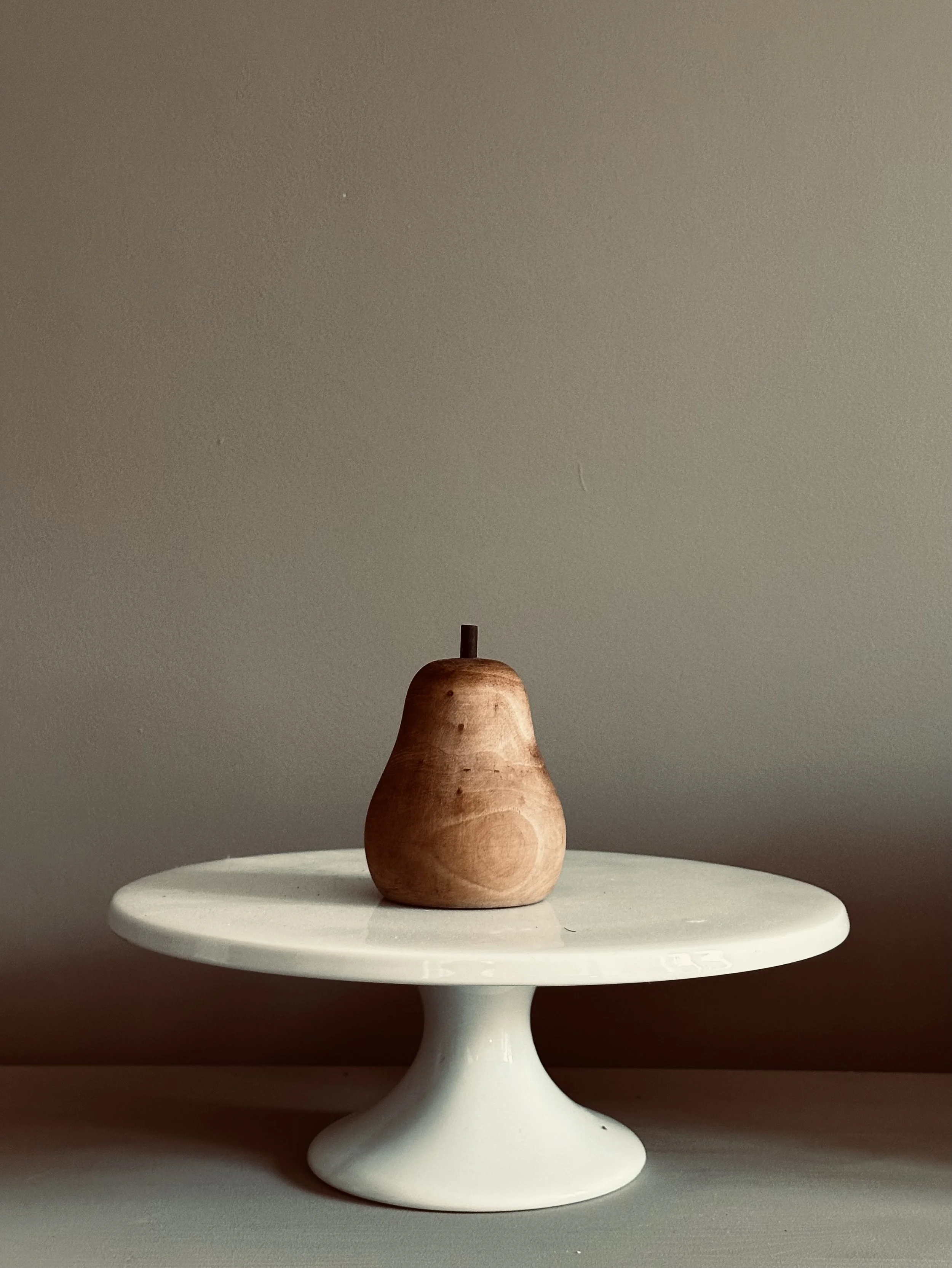 Wooden pear-shaped object on a white pedestal cake stand against a neutral background.