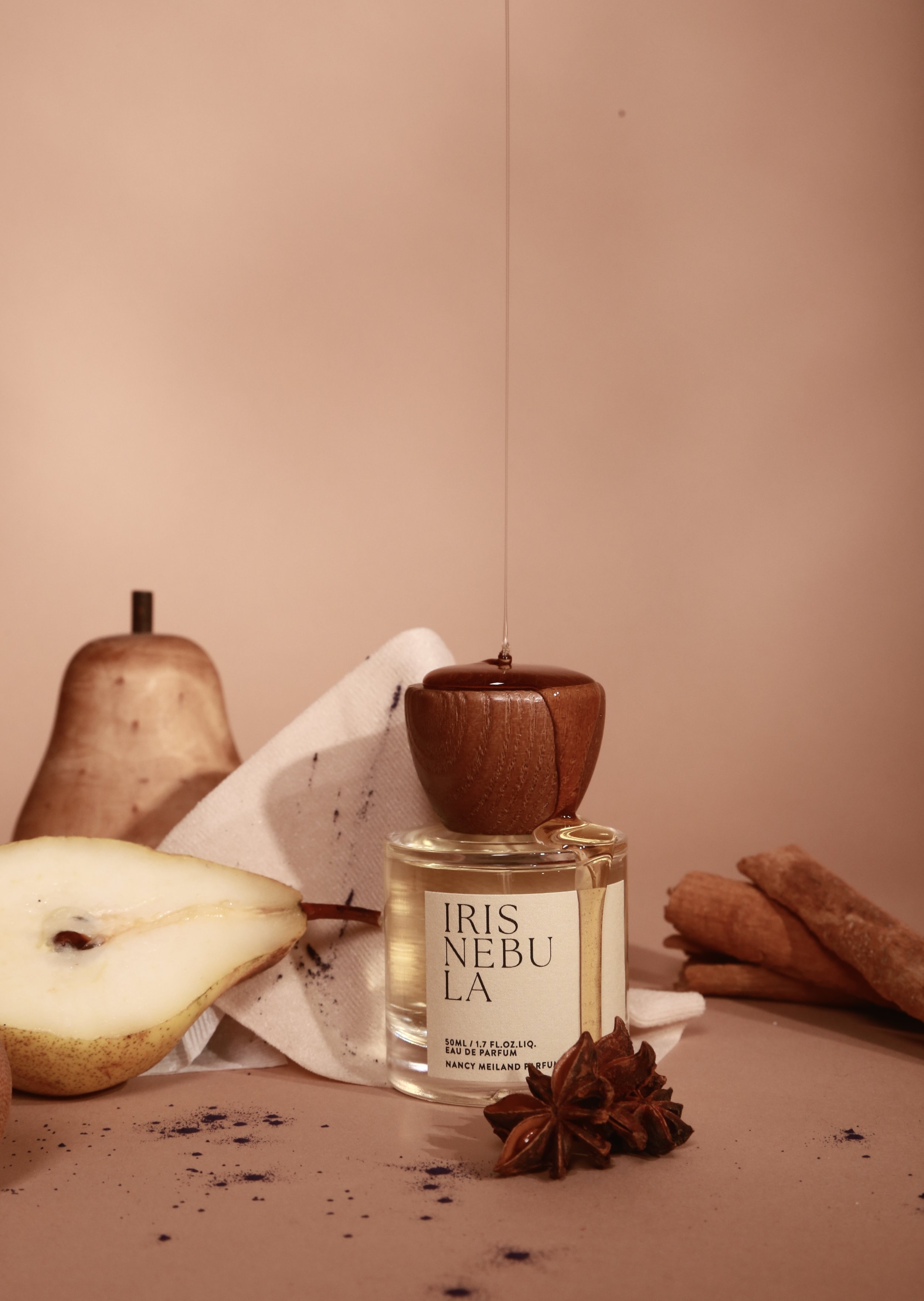 A perfume bottle labeled 'IRIS NEBULA' surrounded by a pear, cinnamon sticks, star anise, and a cloth, with a soft pink background.