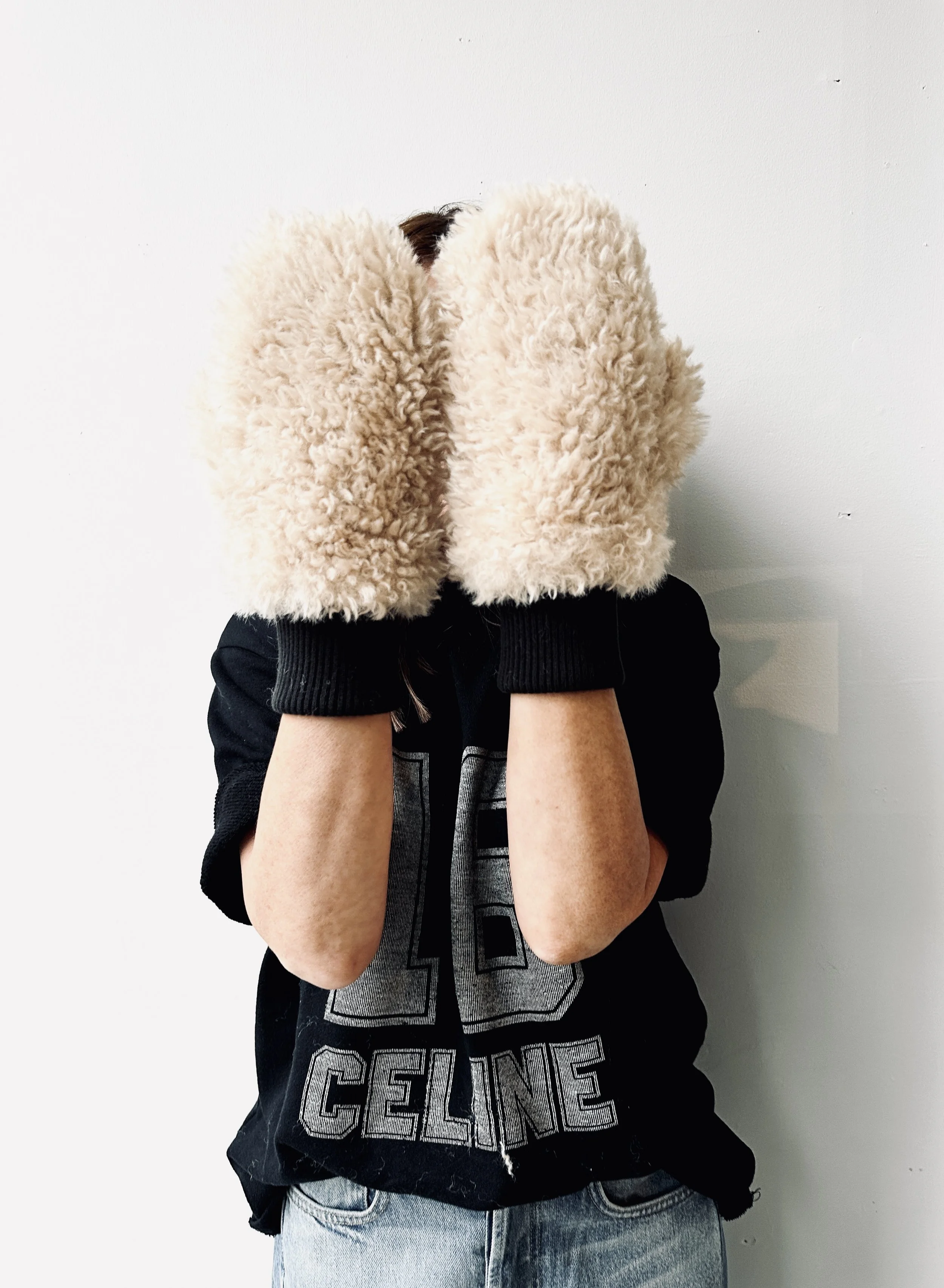 Person wearing large furry beige mittens covering their face, black jacket, and light blue jeans, standing against a plain white wall.
