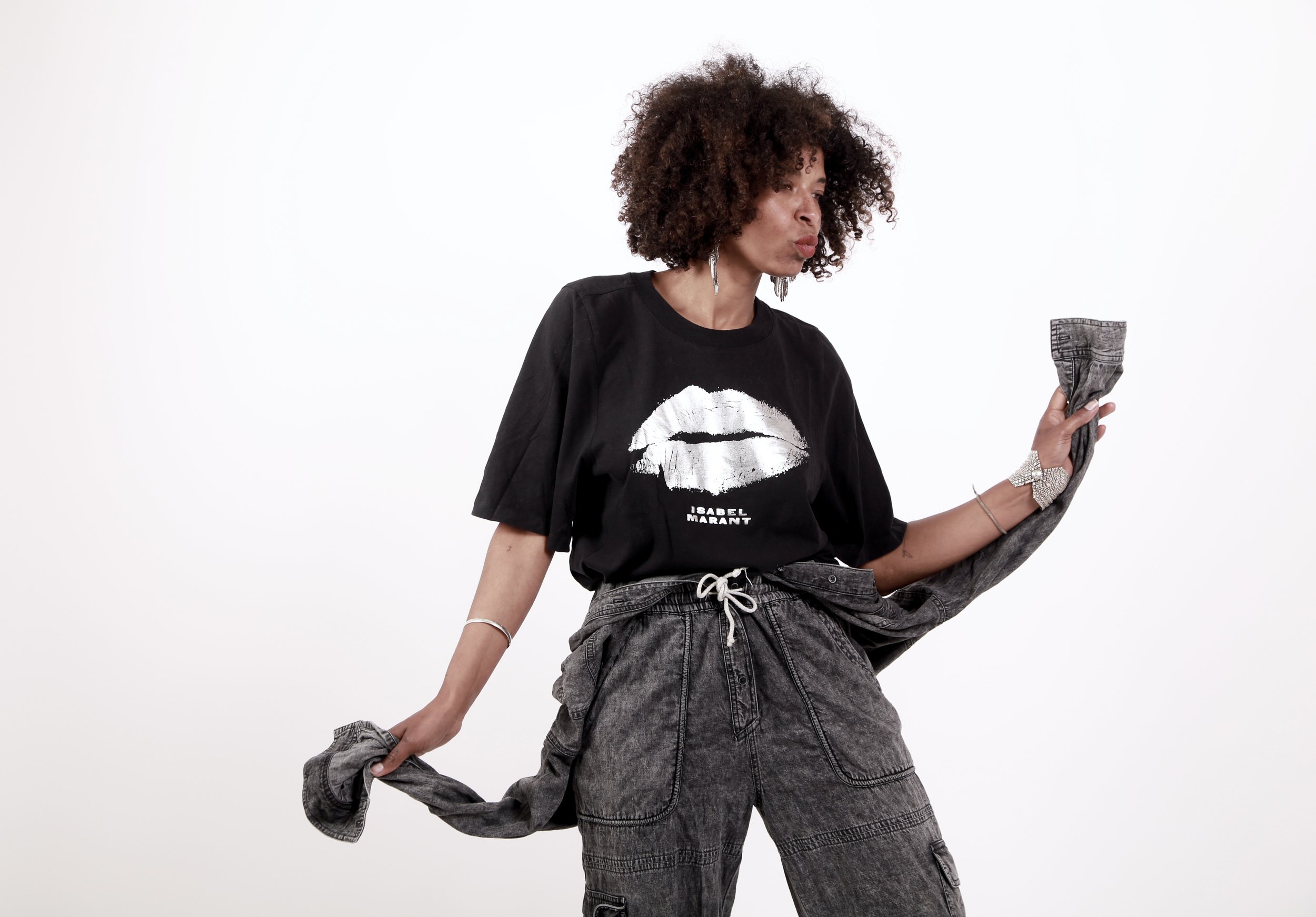 A woman with curly hair wearing a black t-shirt with a white lip graphic and text that says 'Isabel Marant', gray cargo pants, and jewelry, holding a pair of jeans in a fashion pose against a plain white background.
