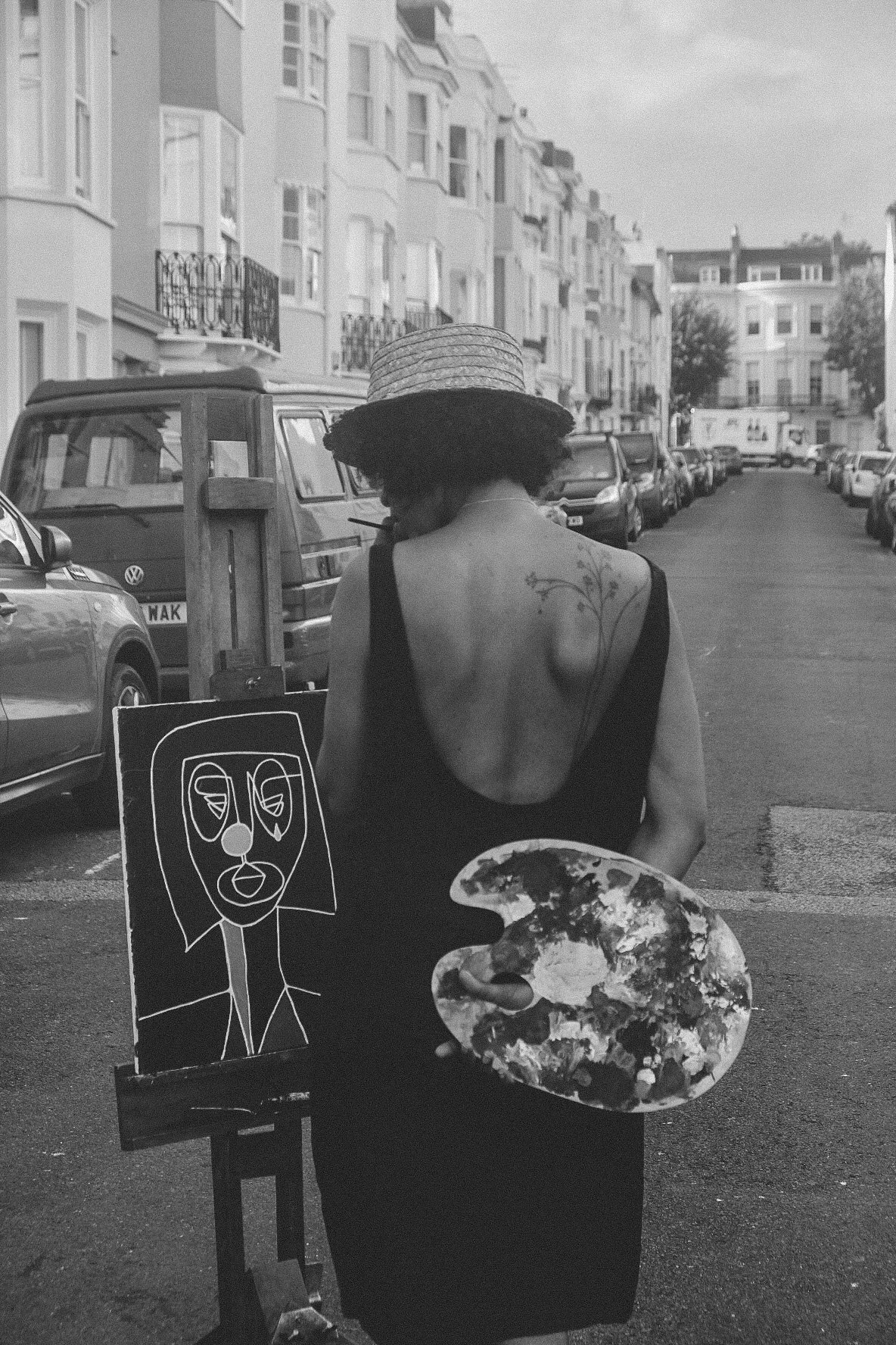 A woman with curly hair wearing a wide-brimmed straw hat and a backless black dress holding an artist's palette, standing in a city street with parked cars and residential buildings in the background.