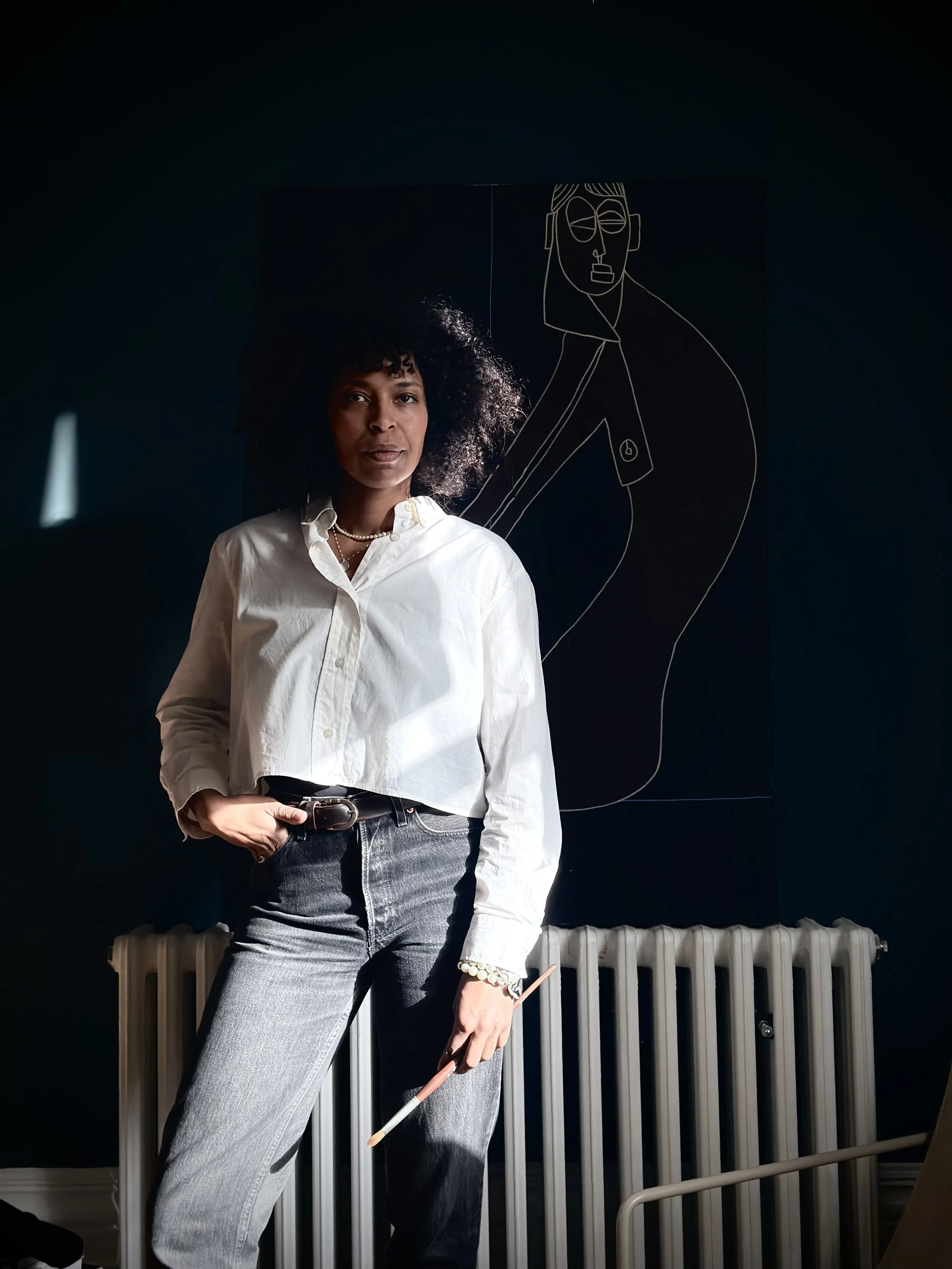 A woman with curly hair standing indoors, wearing a white button-up shirt and gray jeans, holding a paintbrush, with a dark background and a line art drawing of a face behind her.