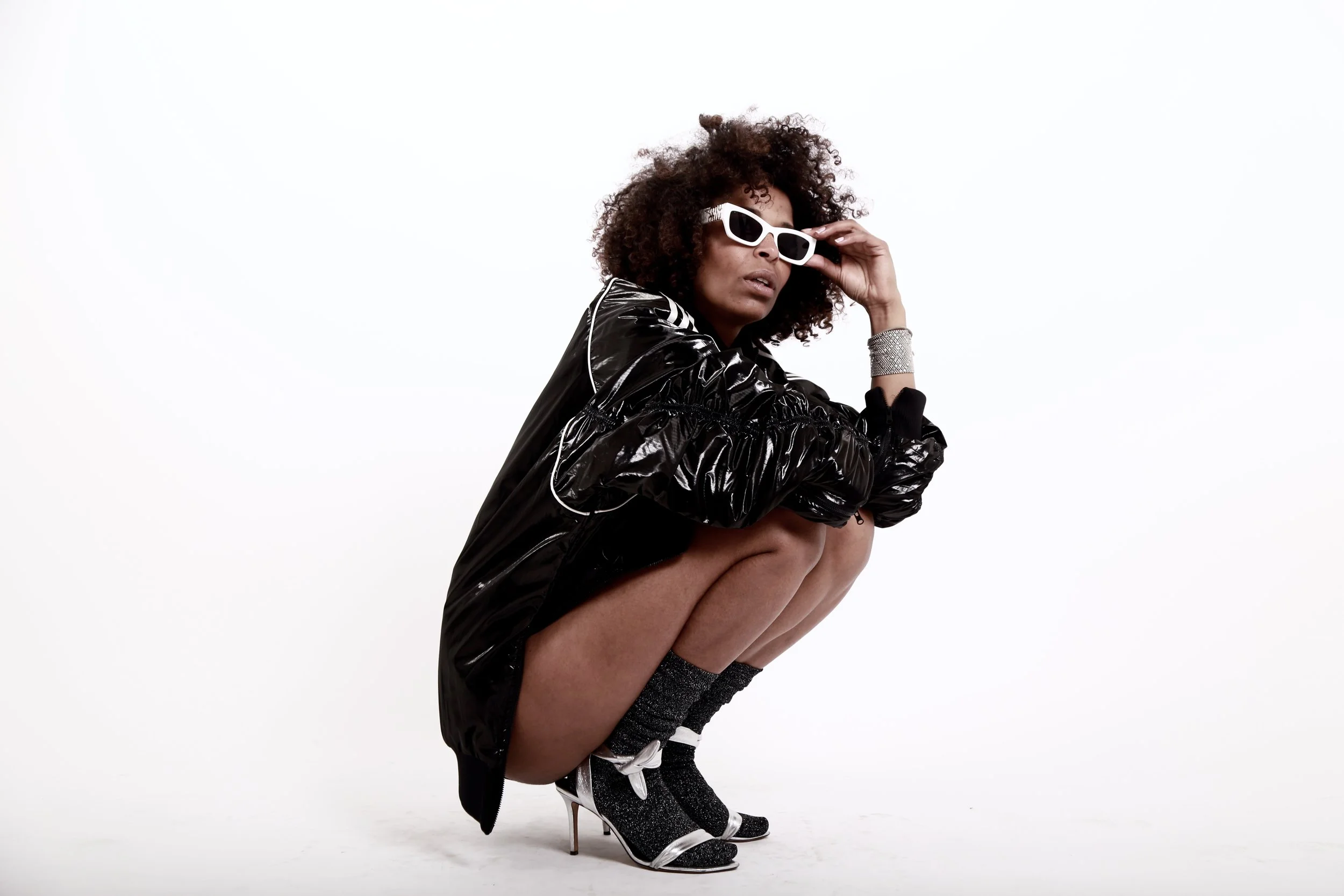 Woman squatting in stylish black jacket, white sunglasses, and high-heeled shoes with socks, against a plain white background.