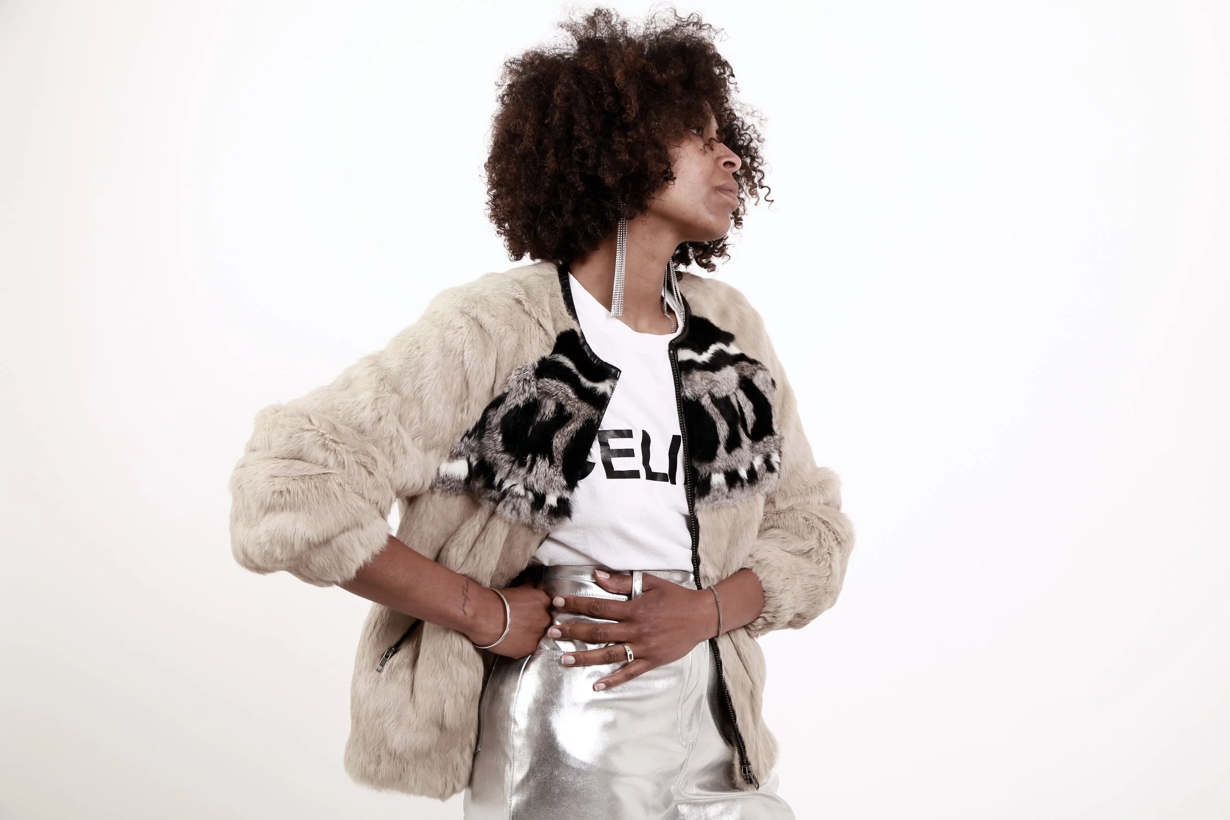 Woman with curly hair wearing a fur jacket, graphic t-shirt, shiny pants, and long earrings posing against a white background.
