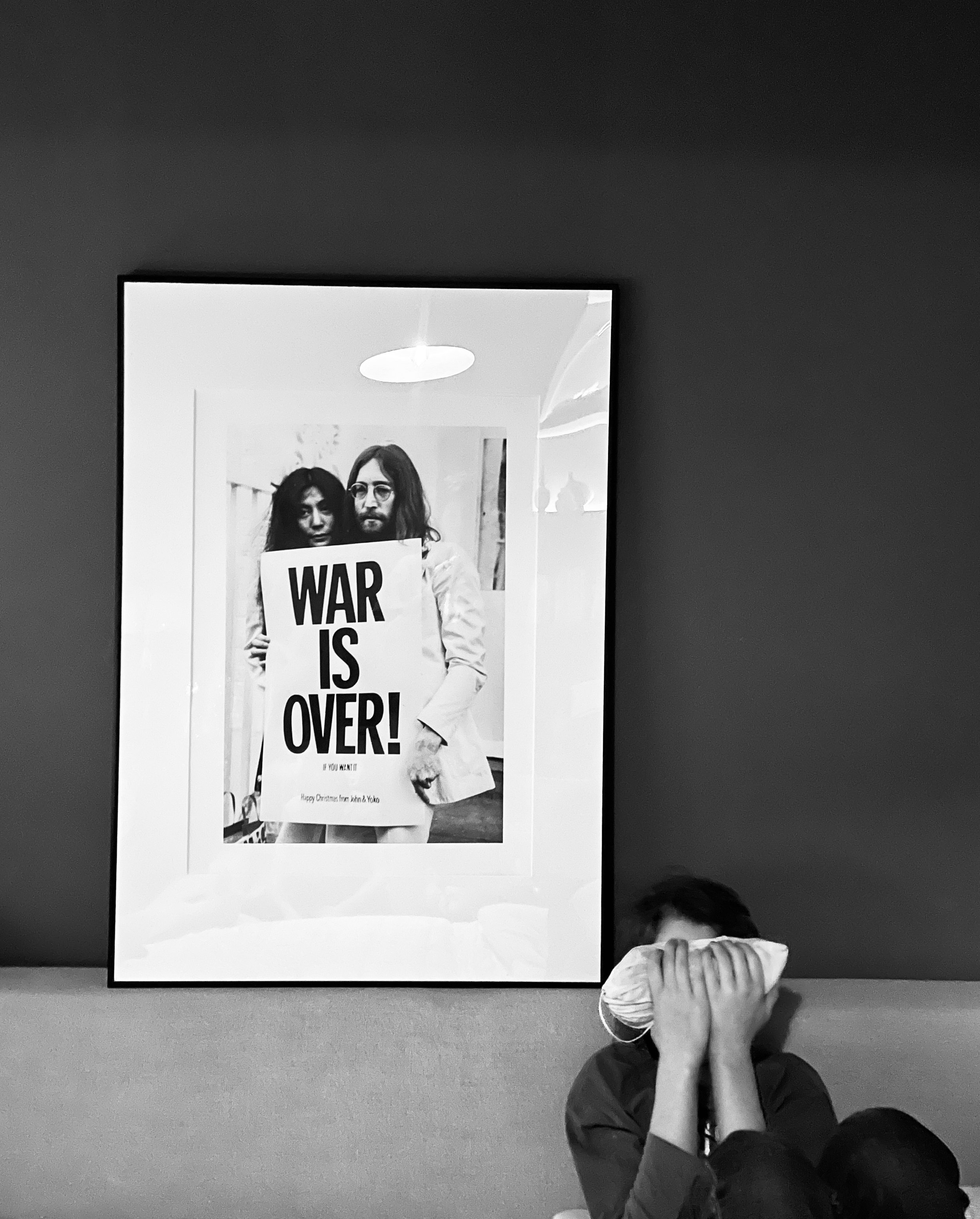 Child sitting on a couch covering their face with a mask in front of a wall with a large black-and-white photograph of two people holding a sign that says 'WAR IS OVER!'