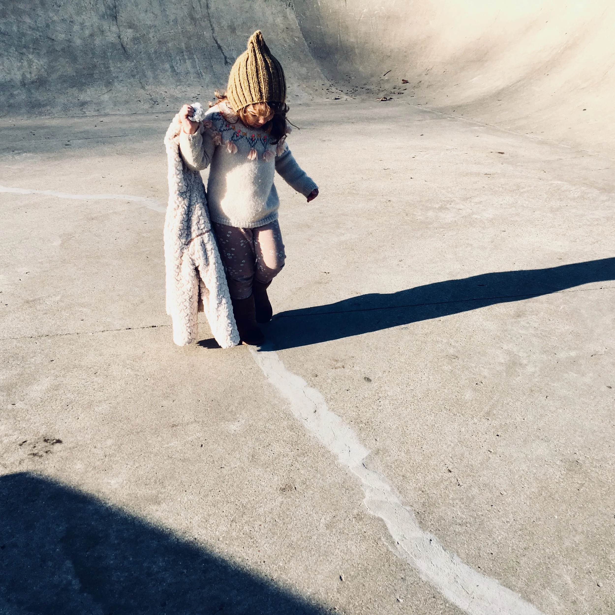 A young girl wearing a brown knit hat, a cozy sweater with decorative elements, and pink pants stands at the edge of a skatepark bowl, drawing a white chalk line on the concrete ground.