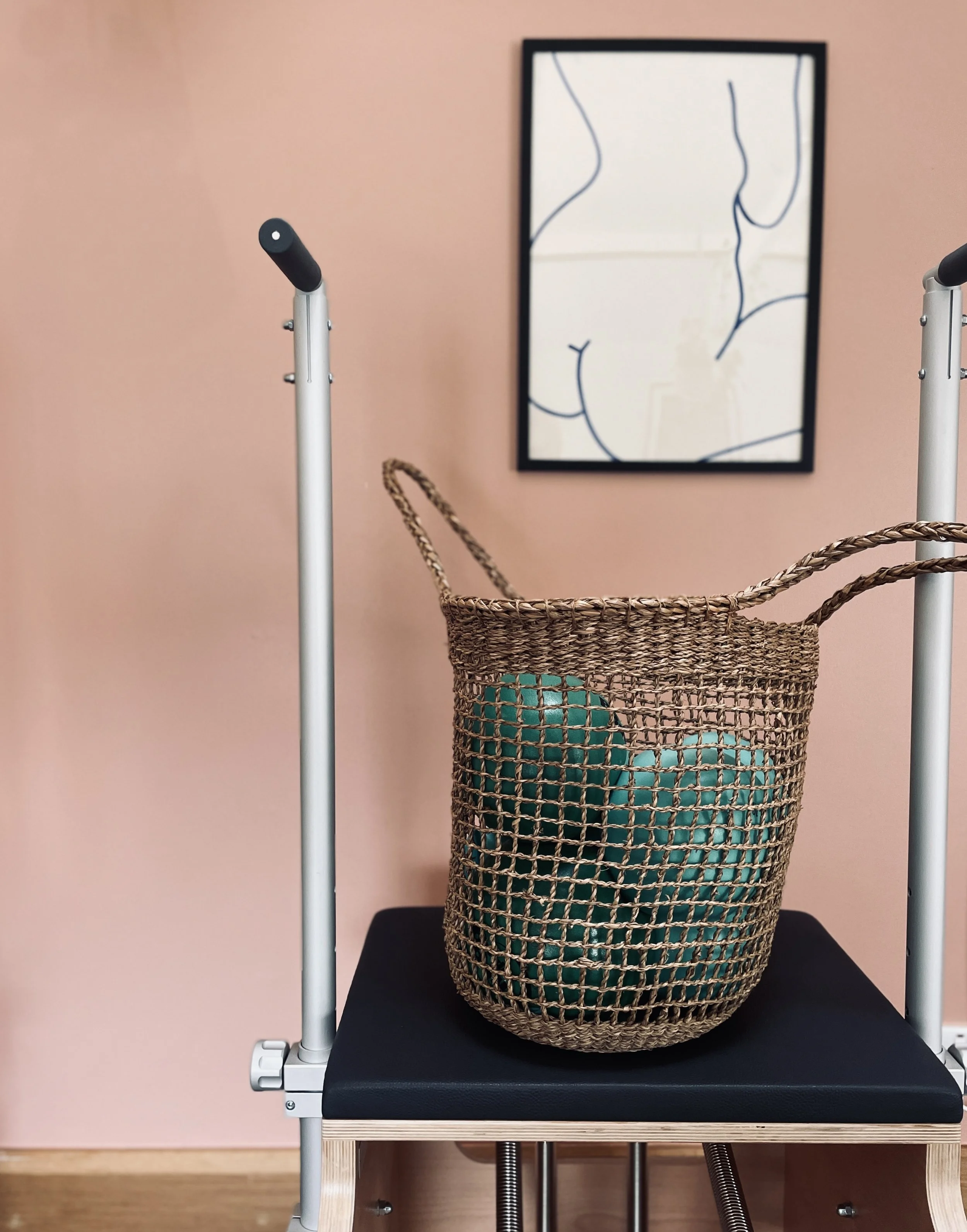A biopsy chair with a wicker basket holding blue balls on it, positioned against a pink wall with an abstract line drawing in a black frame.