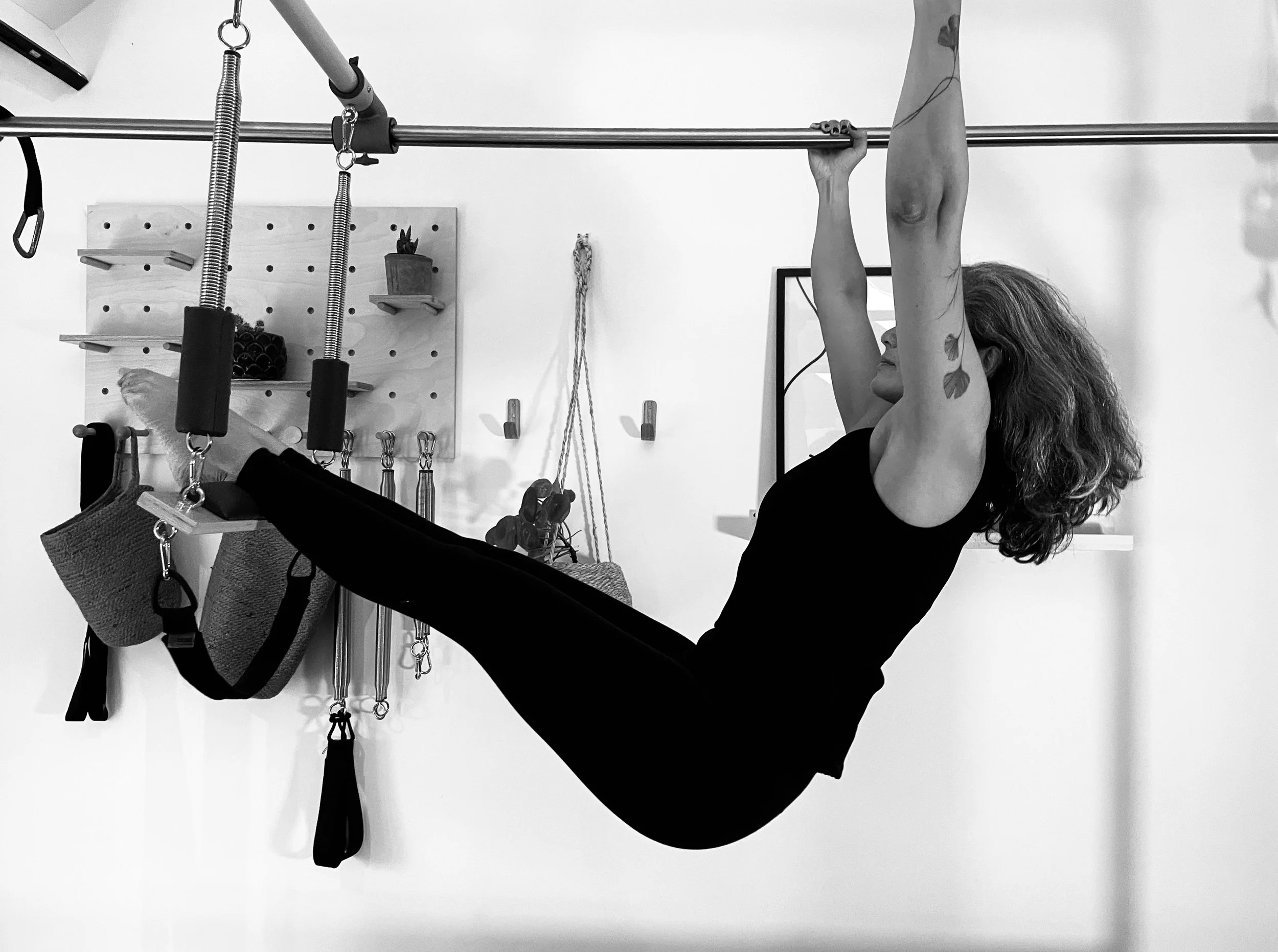 A woman with curly hair hanging upside down from a horizontal bar in a fitness or Pilates studio, using her arms to hold herself up.