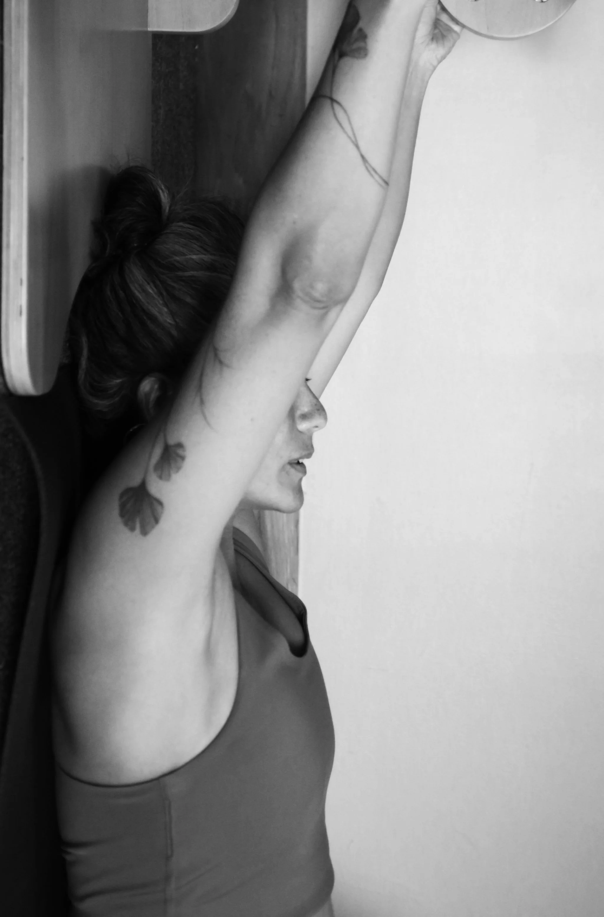 A woman with tattoos on her arm and butterfly earrings is stretching with her arms raised above her head, leaning forward against a table.