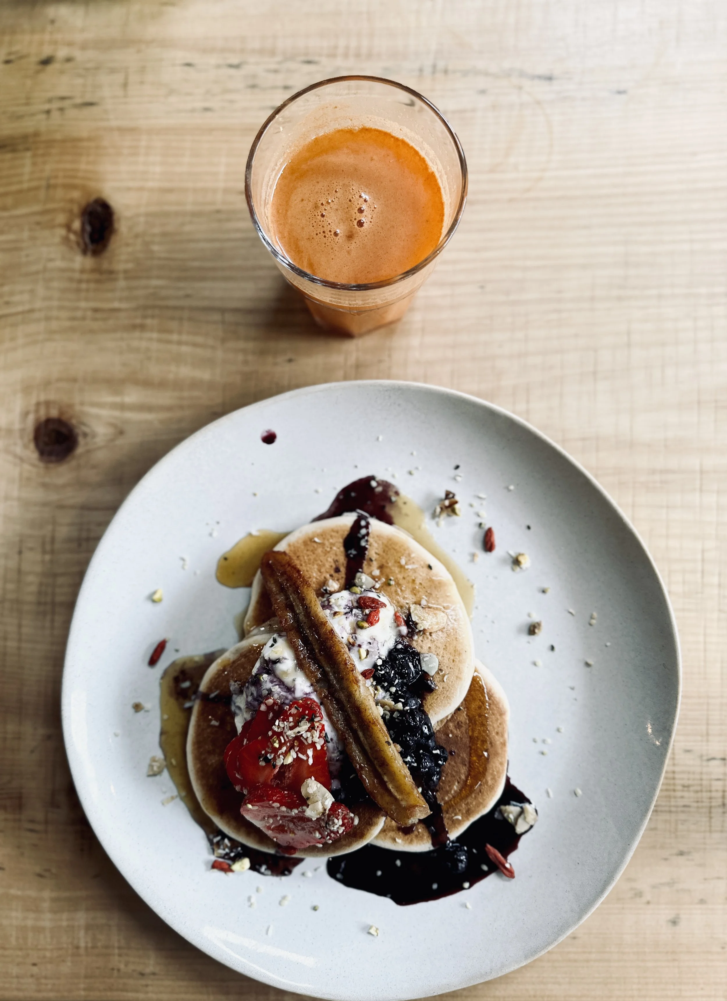 A plate of three pancakes topped with berries, syrup, and nuts, served with a glass of orange juice on a wooden table.