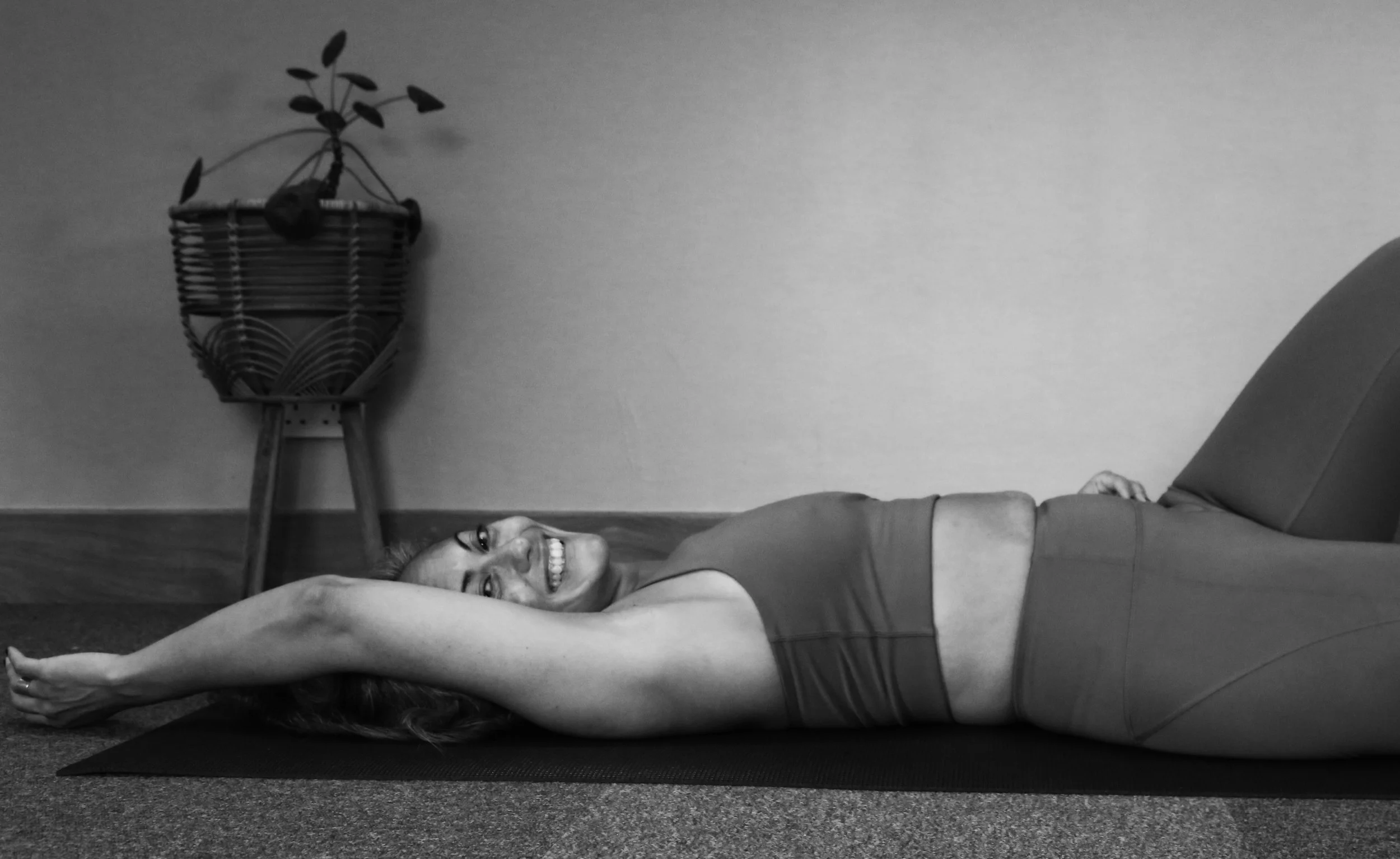 A woman smiling and lying on her back on a yoga mat with her arms extended overhead, in a relaxed pose. She is wearing a sleeveless top and leggings, in a room with a potted plant on a stand in the background.