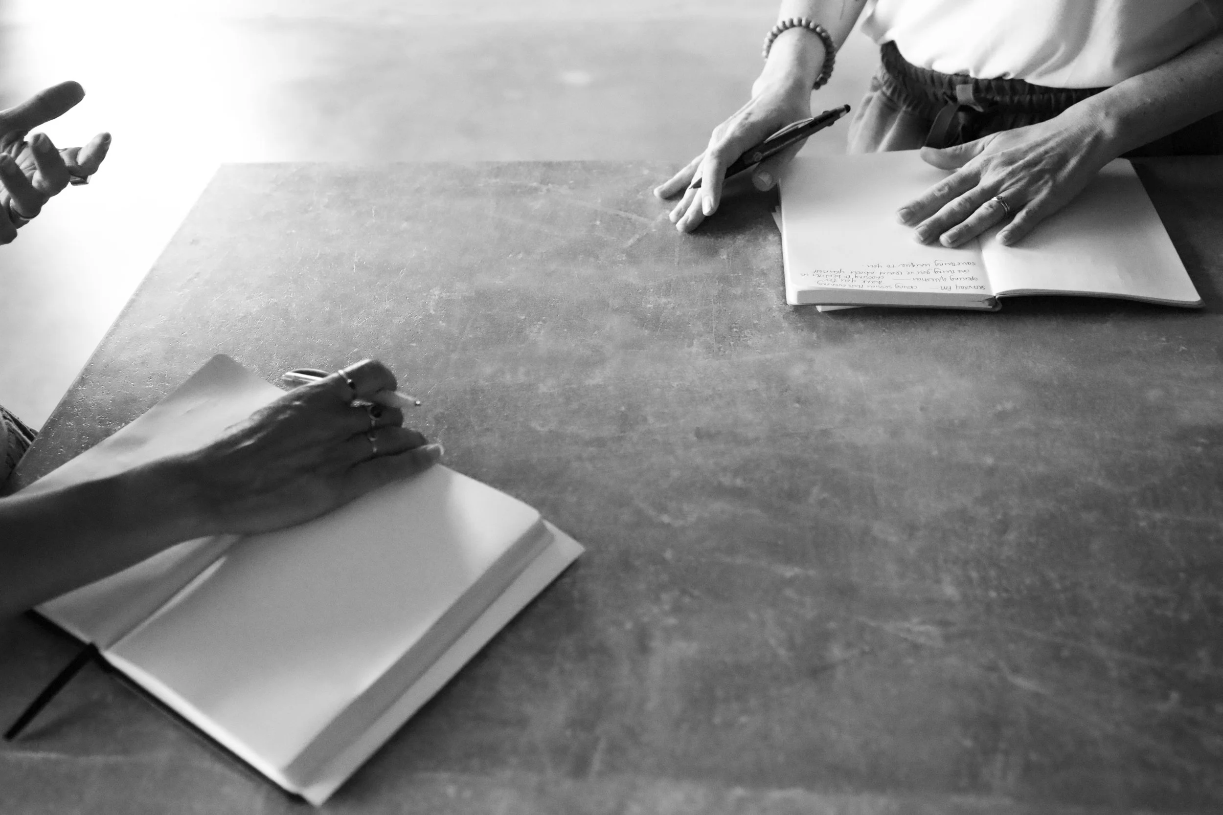 Two people sitting at a table, each with an open notebook, engaging in a discussion or interview, with one person's hand visible on the left and the other person's hands on the right holding a pen and notebook.