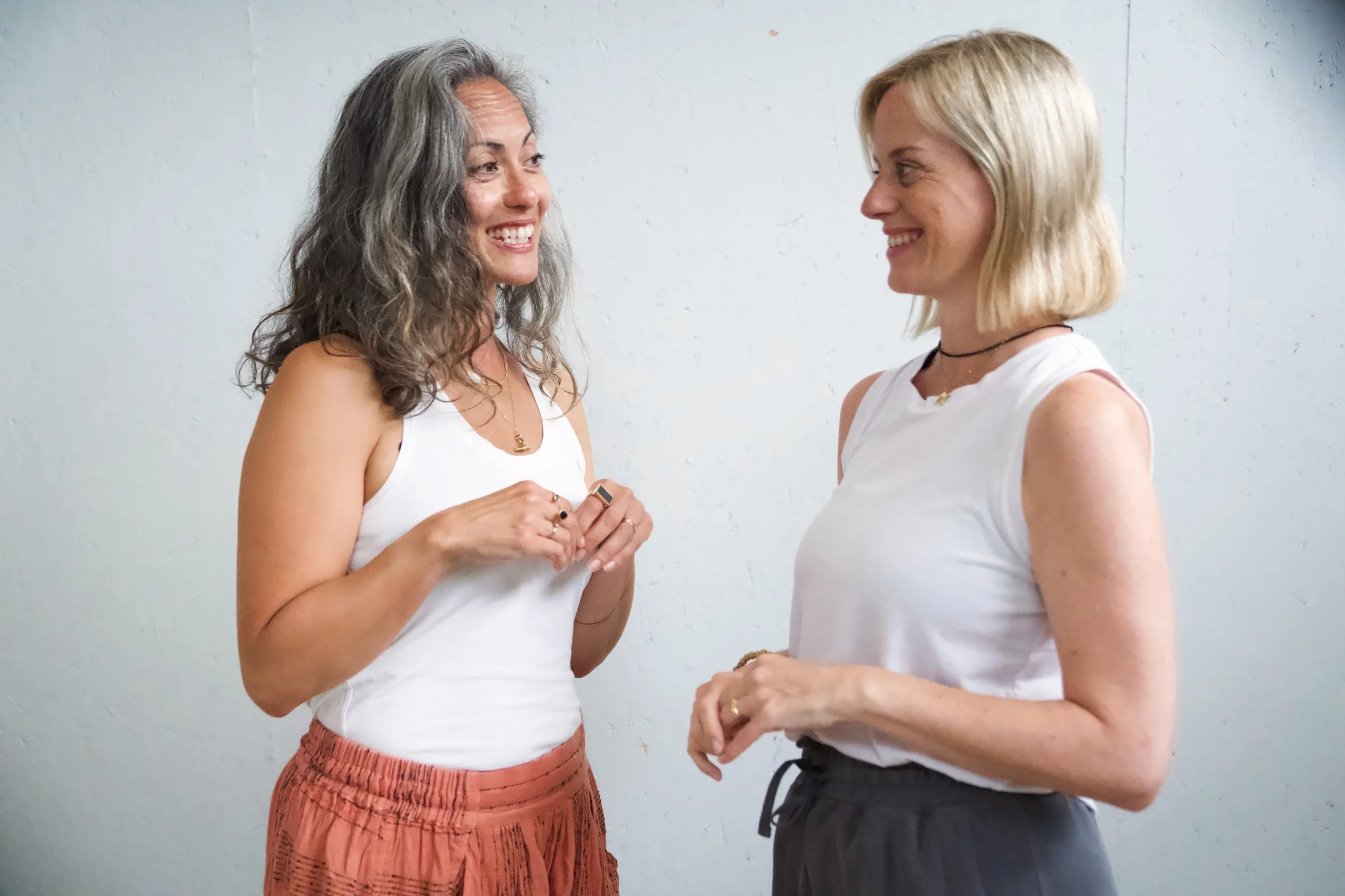 Two women facing each other and smiling, engaged in conversation, wearing casual white tops and dark pants, standing against a plain light-colored wall.