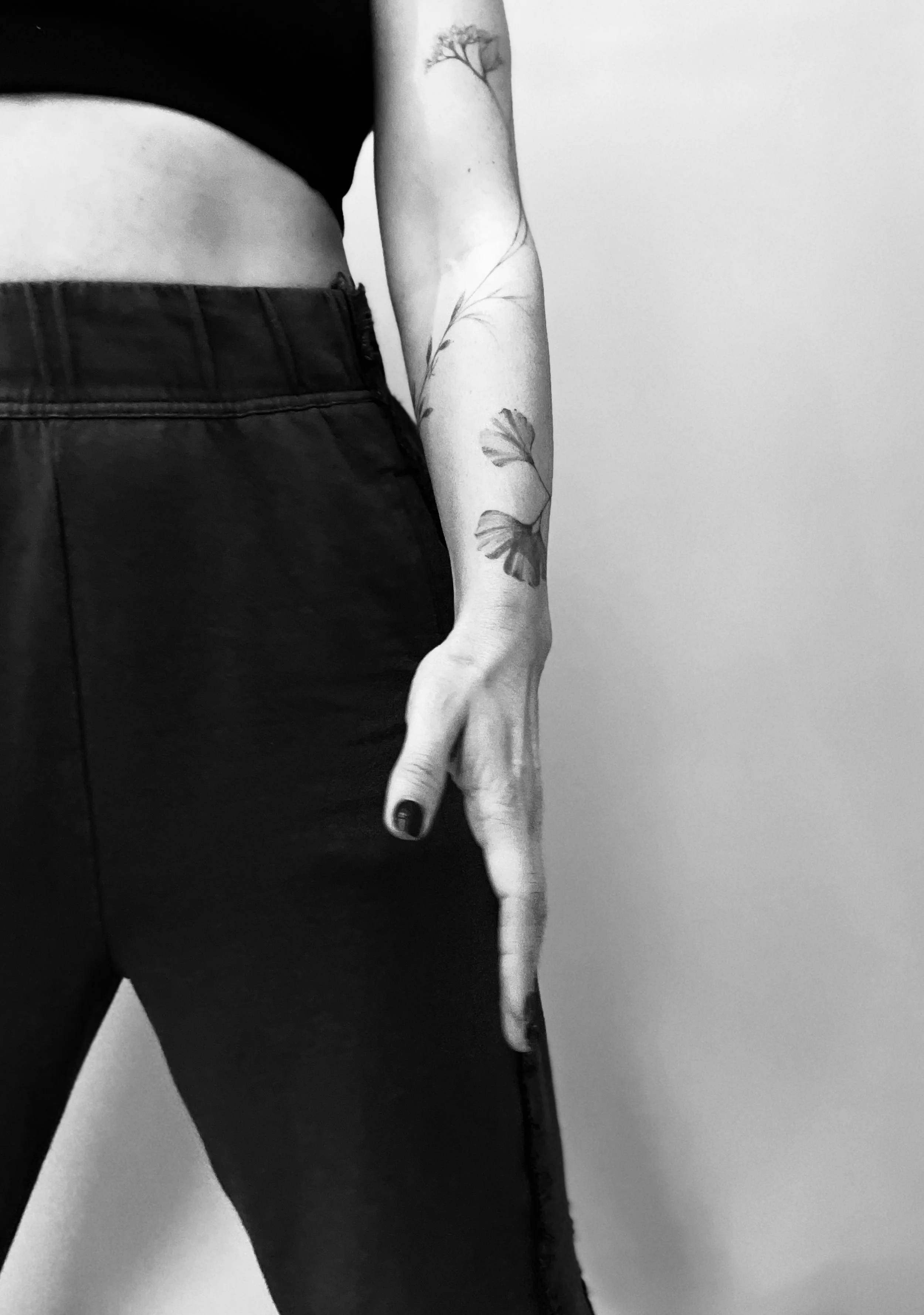 Close-up of a person’s lower torso, arm, and hand, with visible tattoos on the arm, wearing black pants and a black top against a plain background.