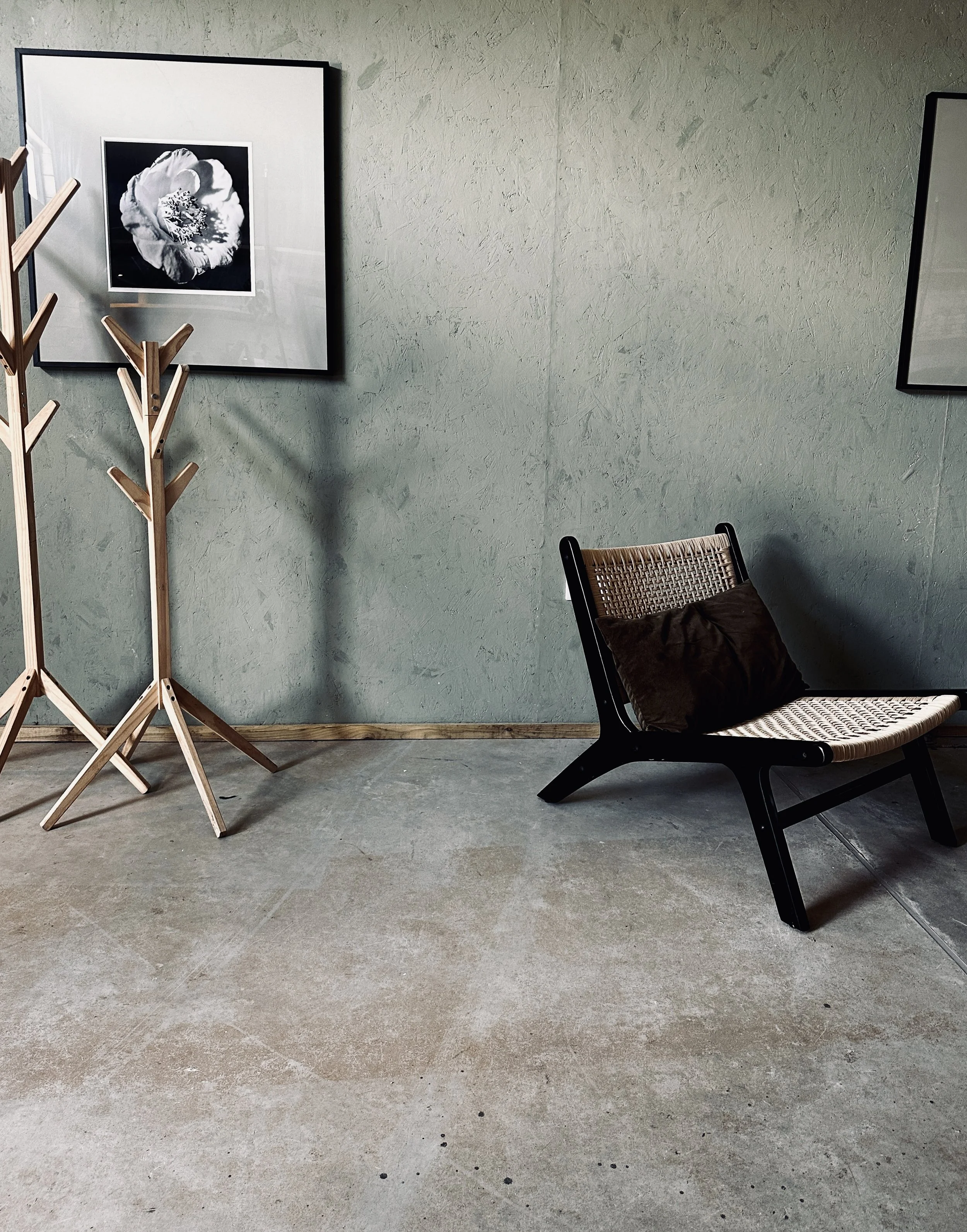 Minimalist room with two wooden coat racks, a black armchair with a brown pillow, and framed black-and-white flower photographs on a textured green wall.