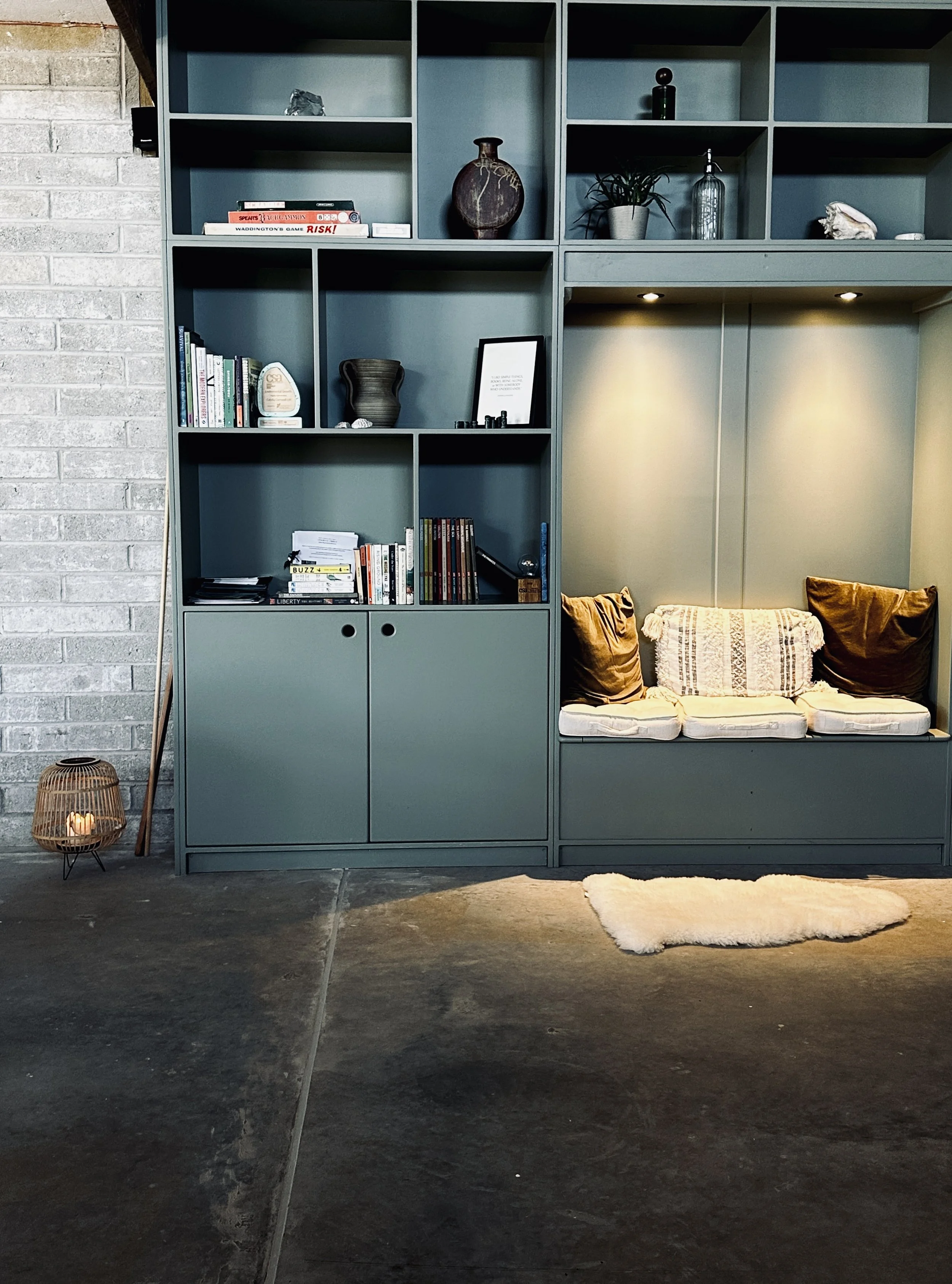 A modern blue bookshelf with decorative items, books, and framed art, next to a cozy built-in seating nook with cushions and pillows, in an industrial-style space with a bookshelf, candle holder, and a white furry rug.