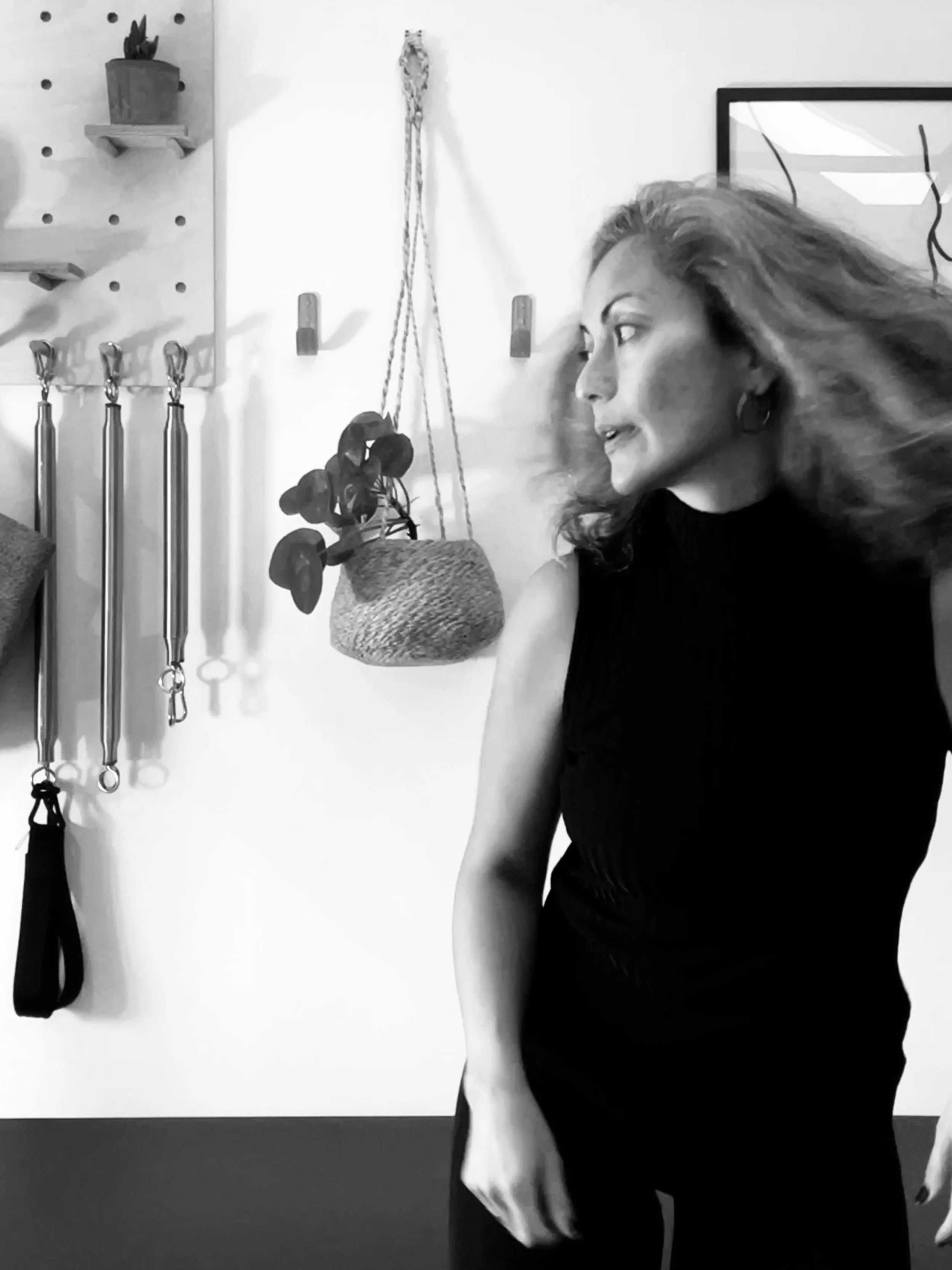 A woman with curly hair wearing a black sleeveless top standing in front of a wall with hanging decor, including a potted plant, ropes, and hooks.