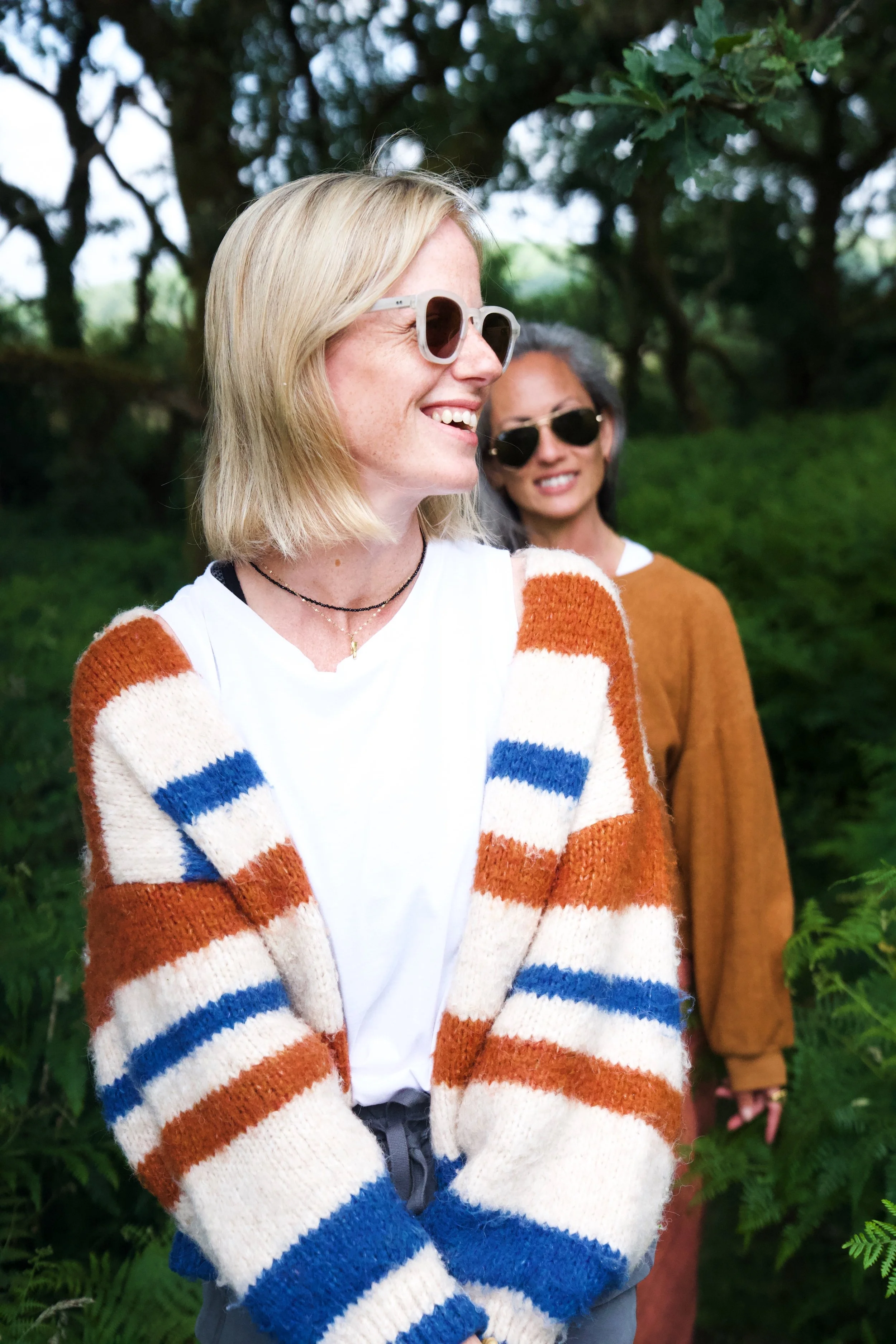 Two women outdoors, smiling, wearing sunglasses, with trees and greenery in the background. The woman in the foreground has blonde hair, freckles, and is wearing a striped cardigan in orange, blue, and white over a white shirt, and a black necklace.