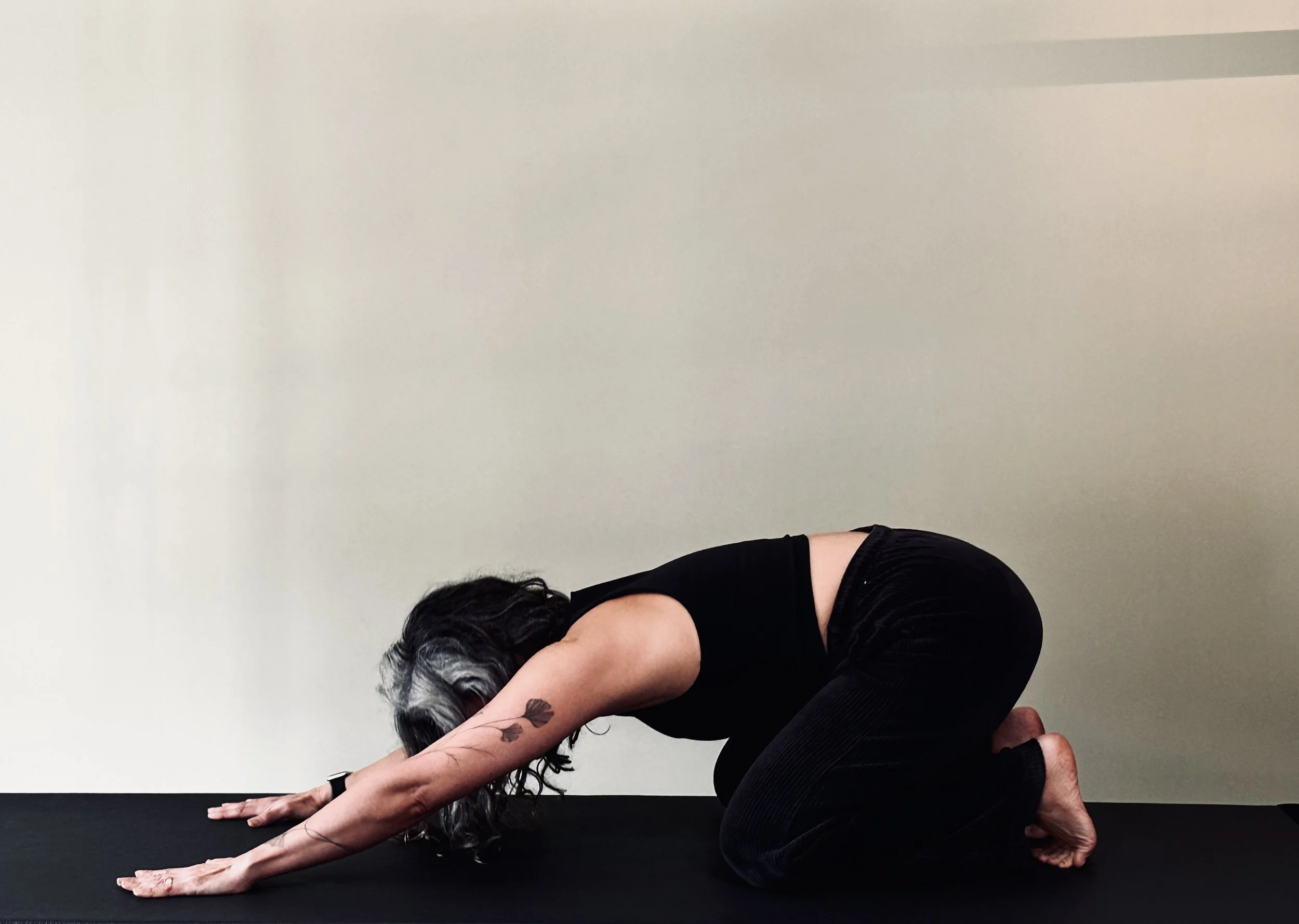 A woman with long dark hair, wearing a black sleeveless top and black pants, practicing yoga on a black mat, in a kneeling position with arms extended forward and head down.