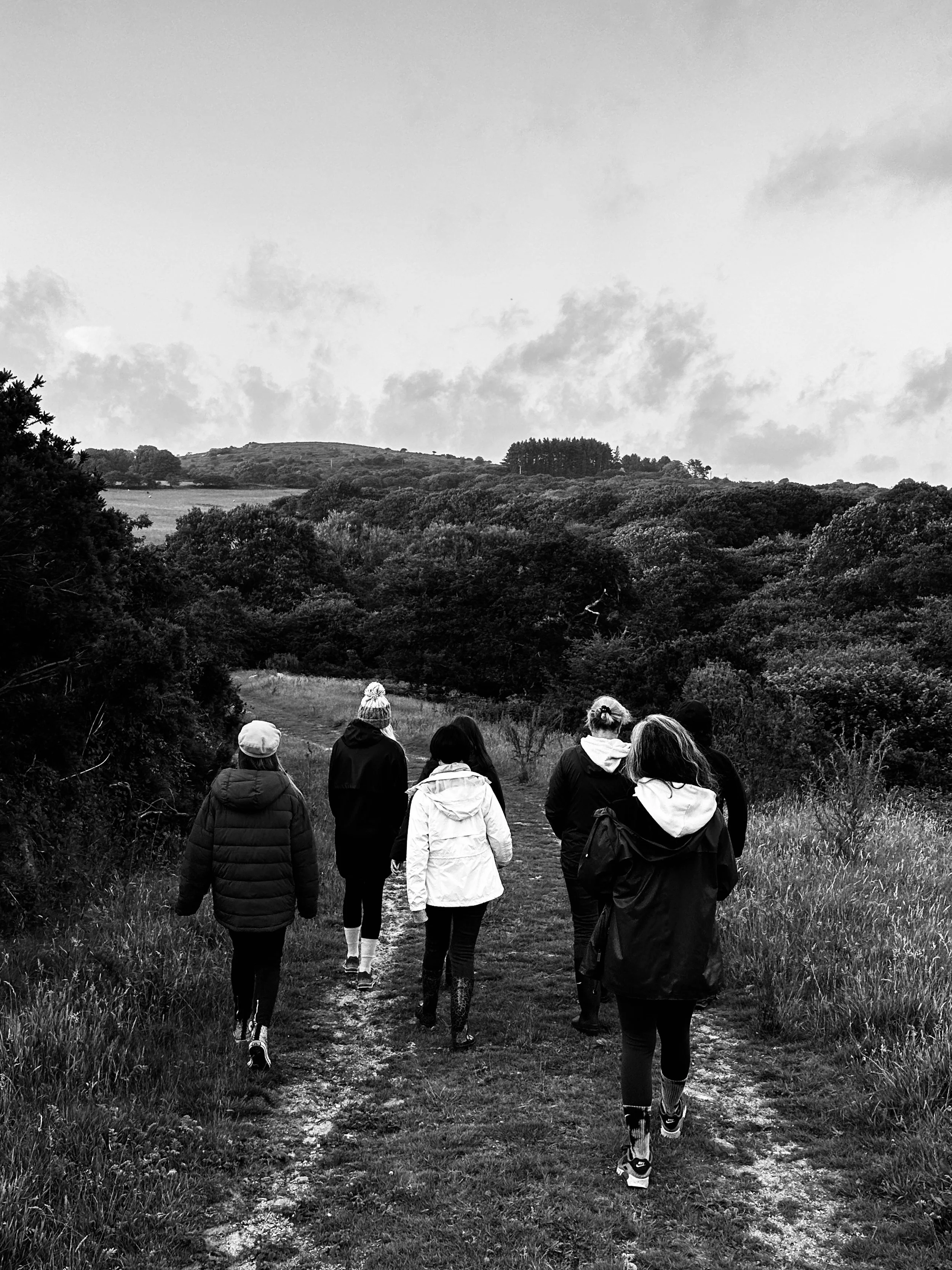 A group of eight people walking on a trail through a grassy, wooded landscape with hills and trees in the background. It's a cloudy day.