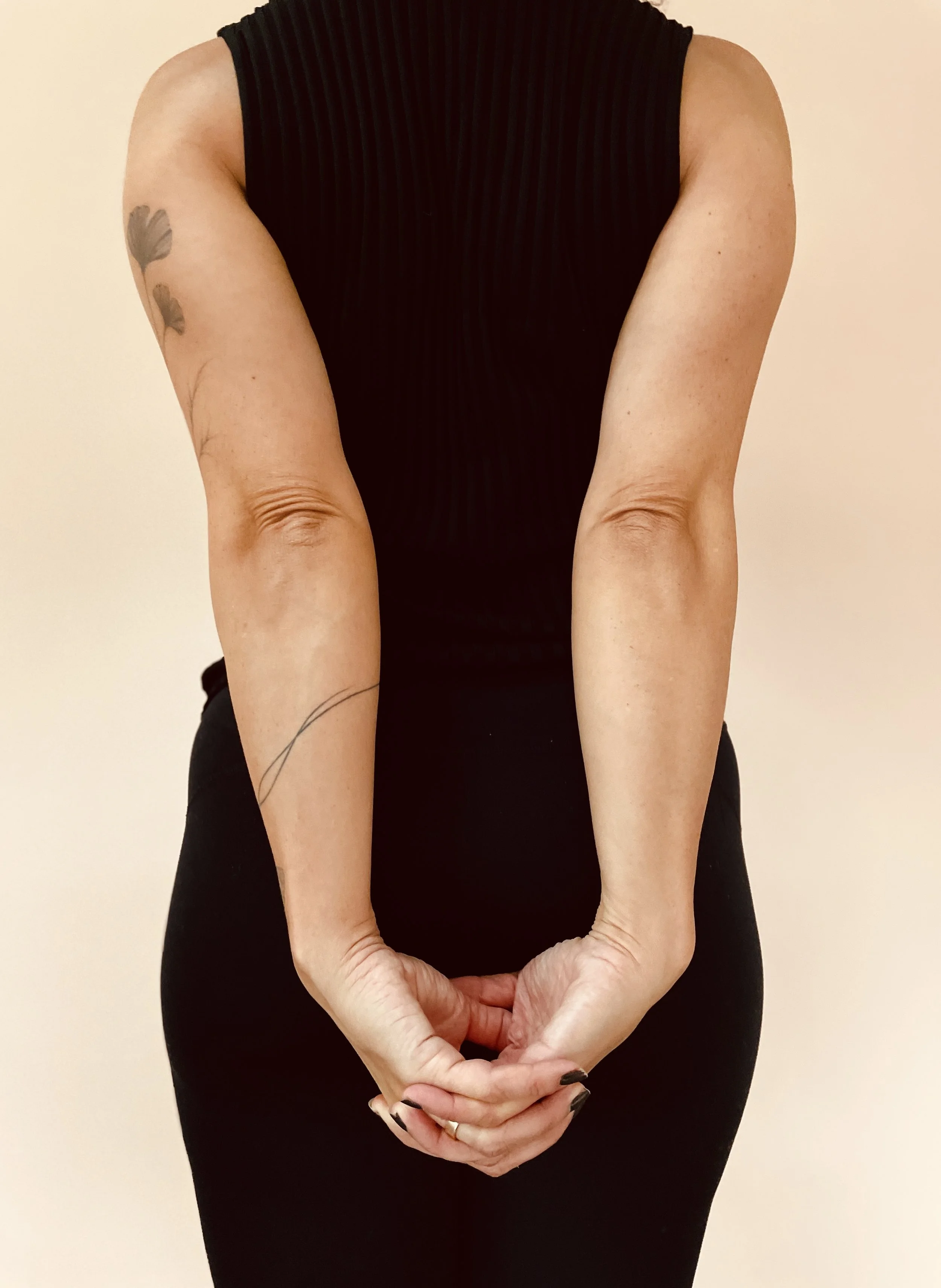 Person in a black sleeveless top with tattoos on both arms, standing against a plain background, with hands lightly clasped behind their back.