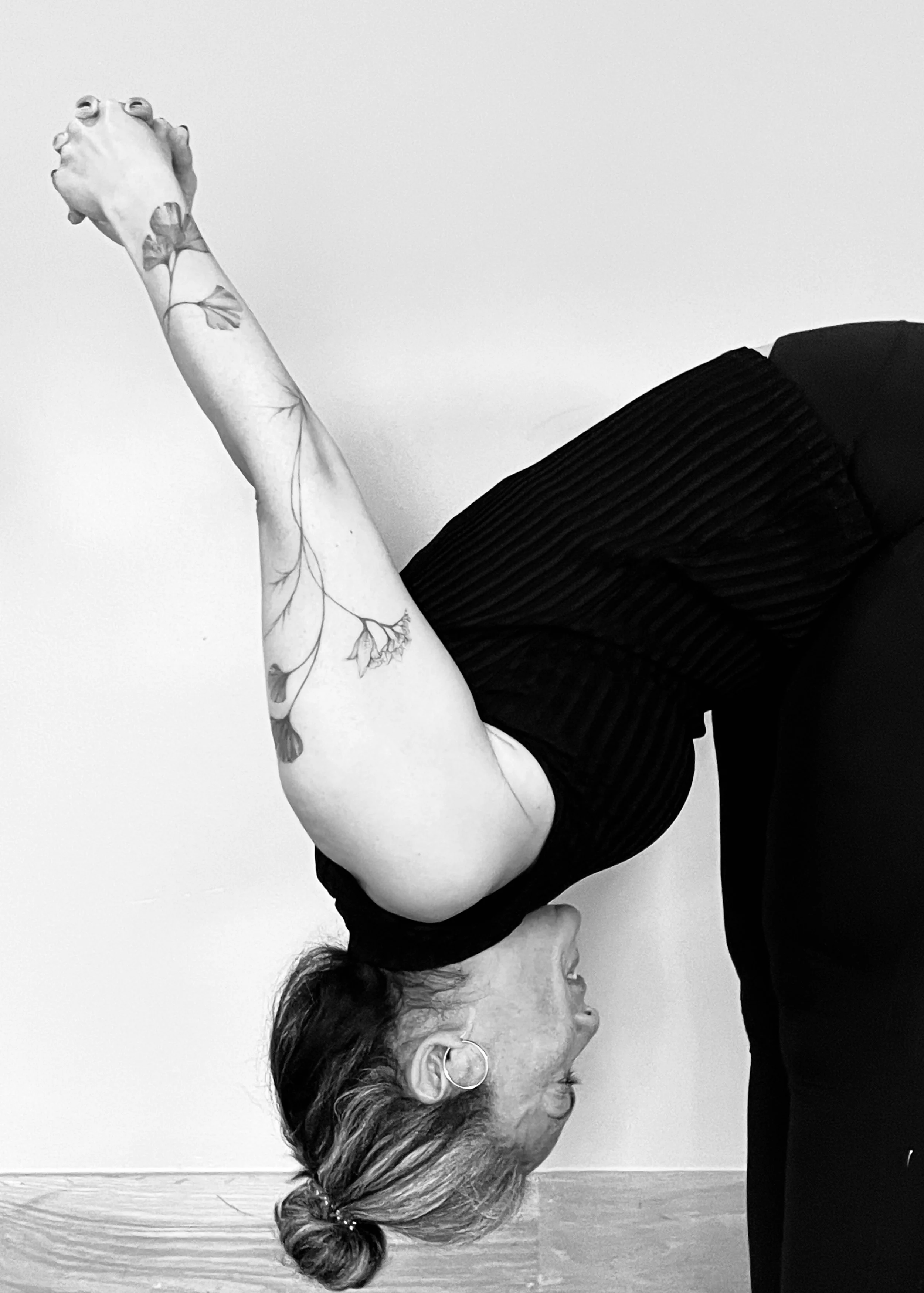 A woman is performing a yoga pose with her body bent forward, one arm extended upward, and her head tilted down. She has tattoos on her arm and is wearing earrings and a black, ribbed sleeveless top.