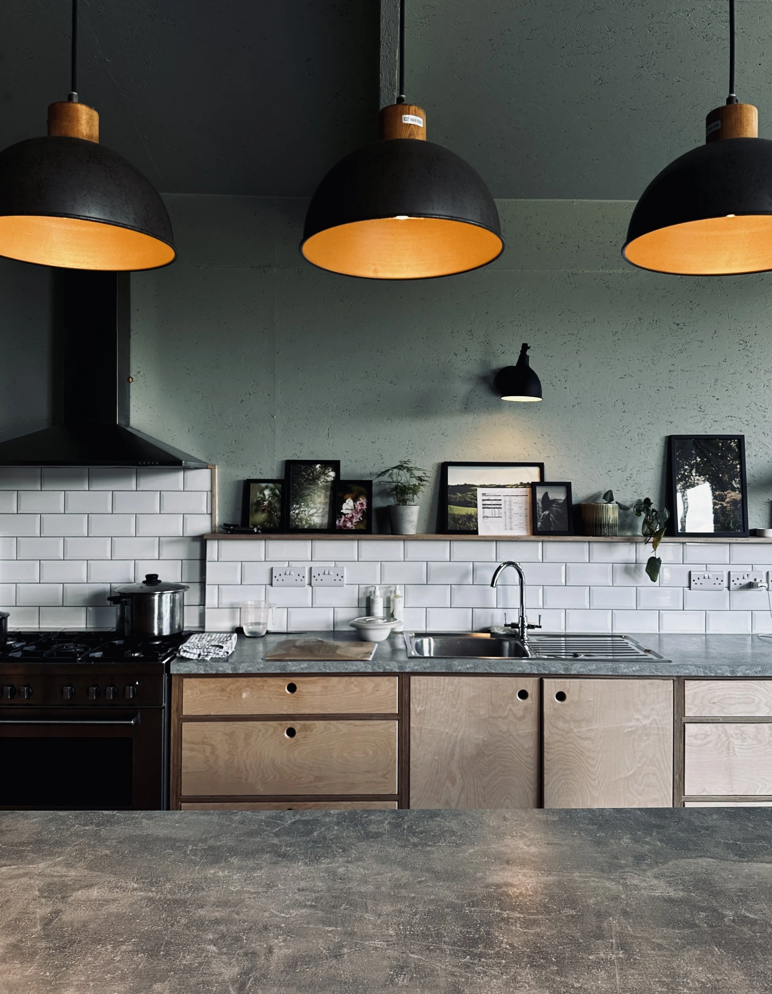 Modern kitchen with black pendant lights, white subway tile backsplash, wooden cabinets, and a countertop with various kitchen items, framed pictures, and potted plants on a shelf.
