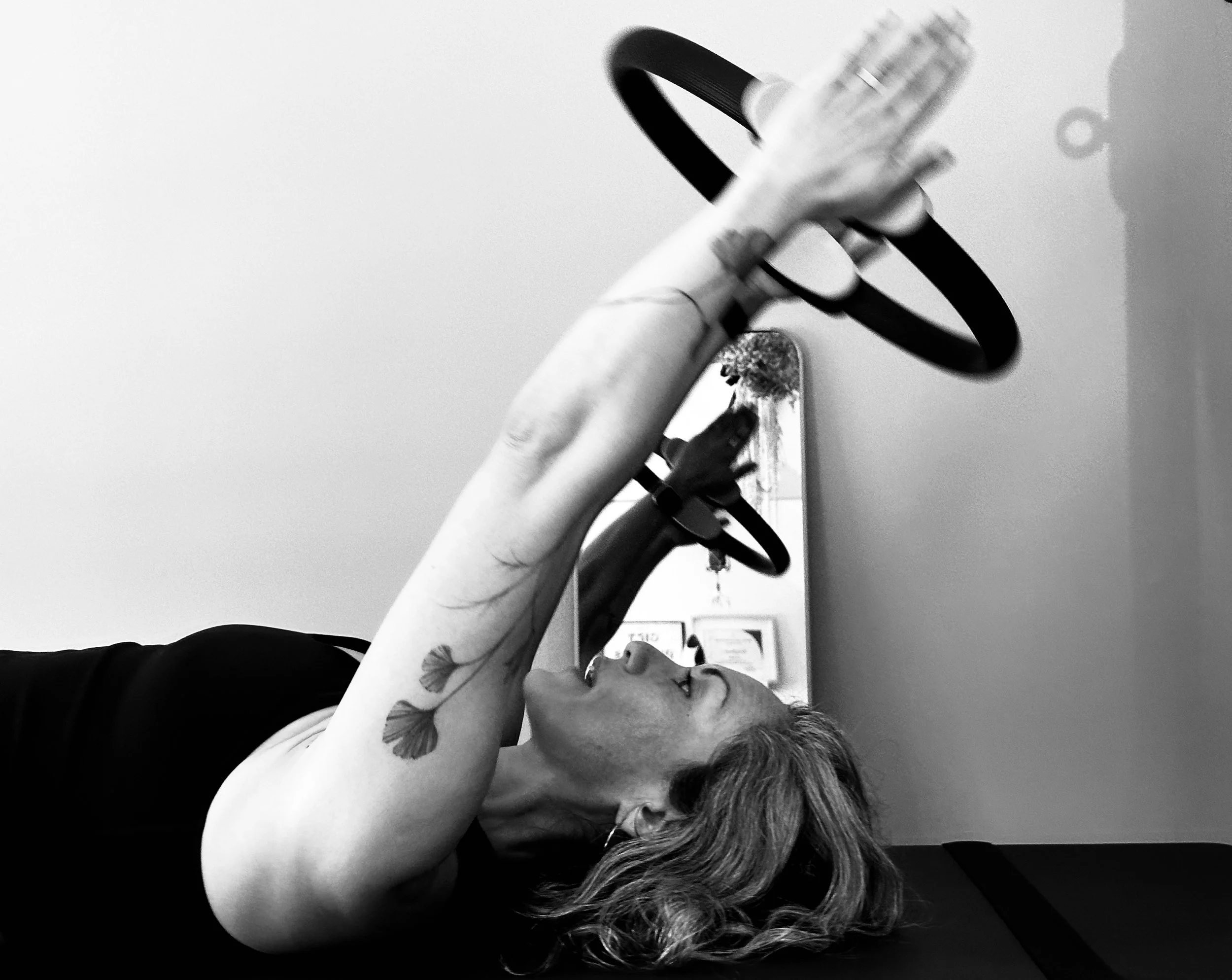 A woman with tattoos on her arm, lying on a surface, holding a fitness ring while looking up at it.