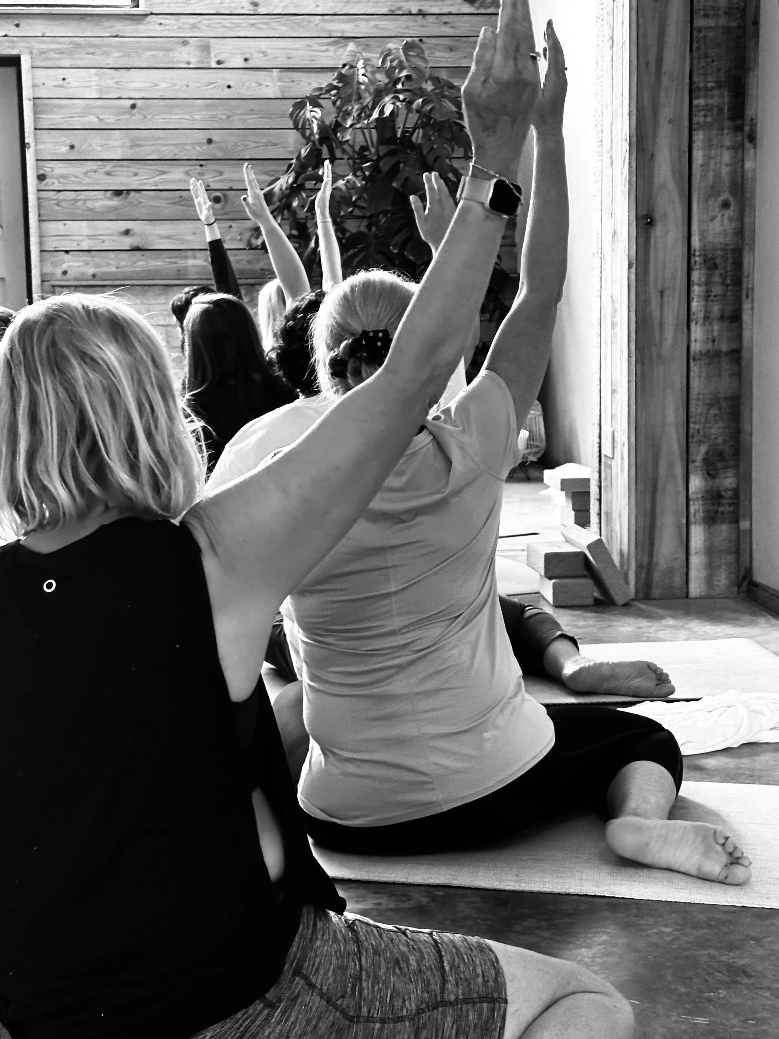 People participating in a yoga or meditation class indoors, sitting on mats with arms raised and eyes closed, with a wooden wall and plant in the background.