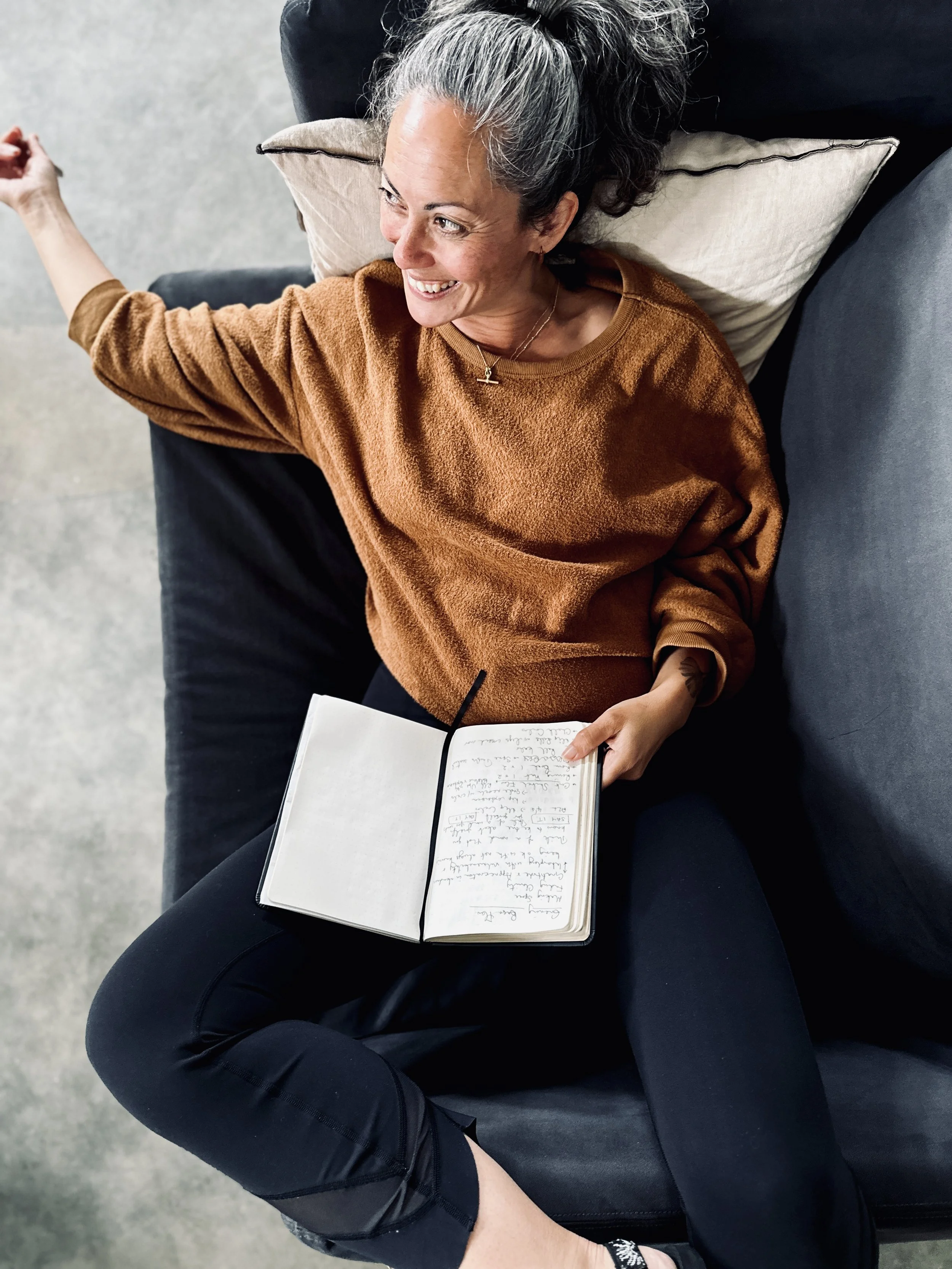 A woman with gray hair lying on a dark sofa, smiling, holding a notebook with handwritten notes, wearing a brown sweatshirt and black pants.