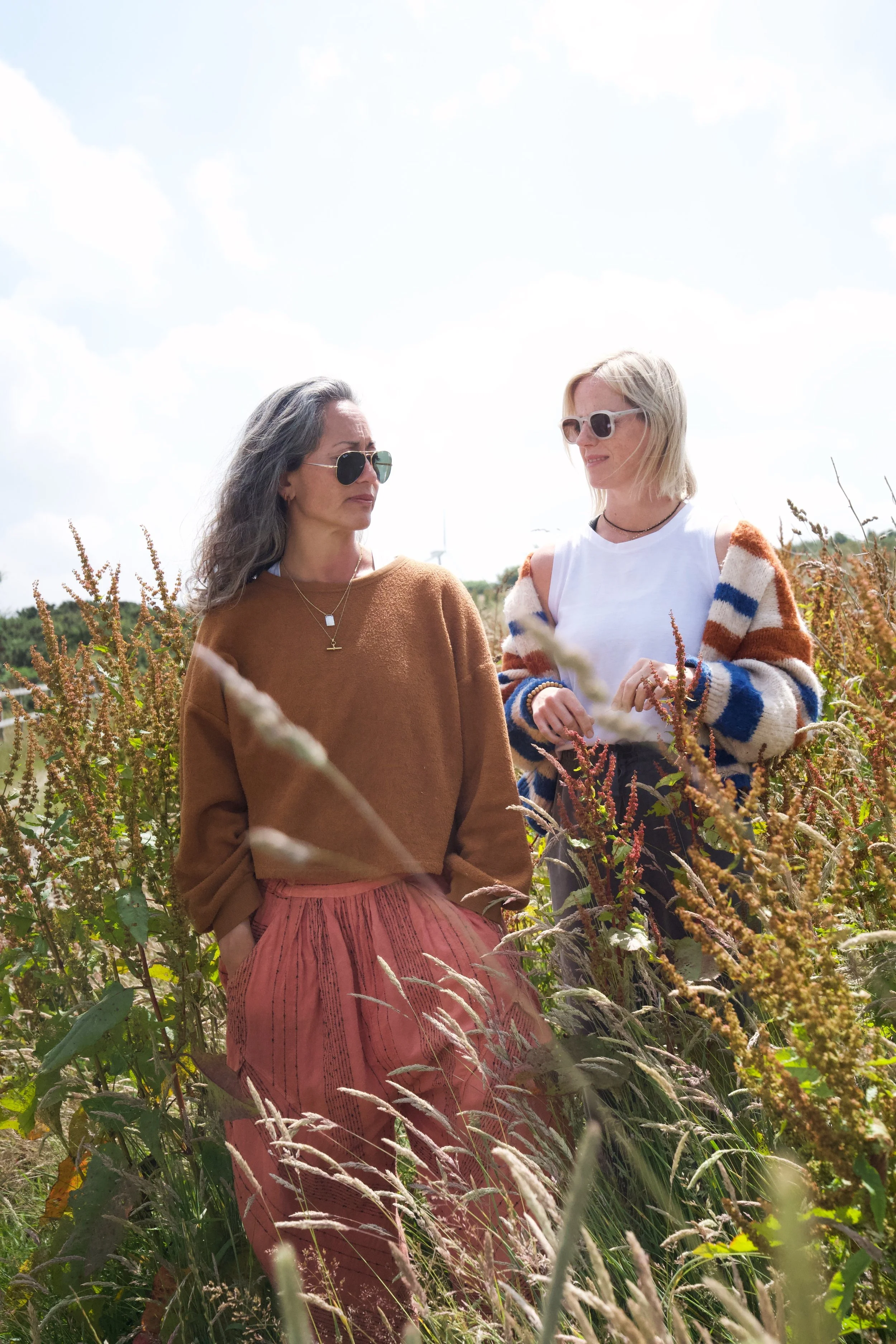 Two women wearing sunglasses standing in a field of tall plants under a cloudy sky.