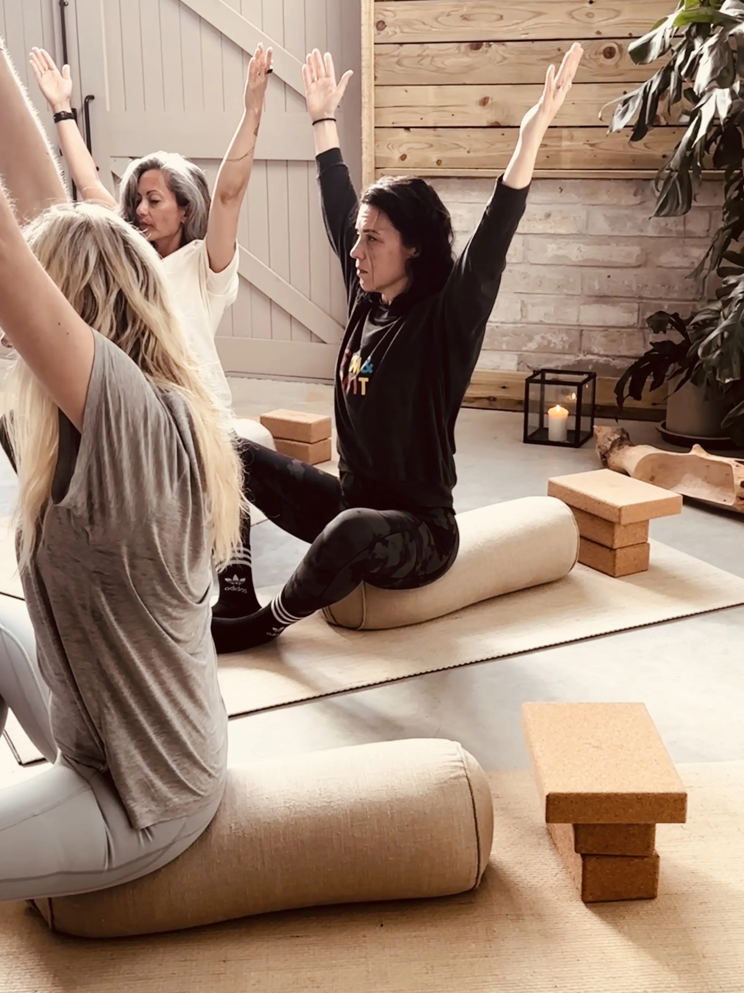 Three women participate in a yoga session in a cozy room with wooden walls, one seated on a bolster with arms raised, others seated nearby in meditation poses, surrounded by candles, plants, and yoga blocks.