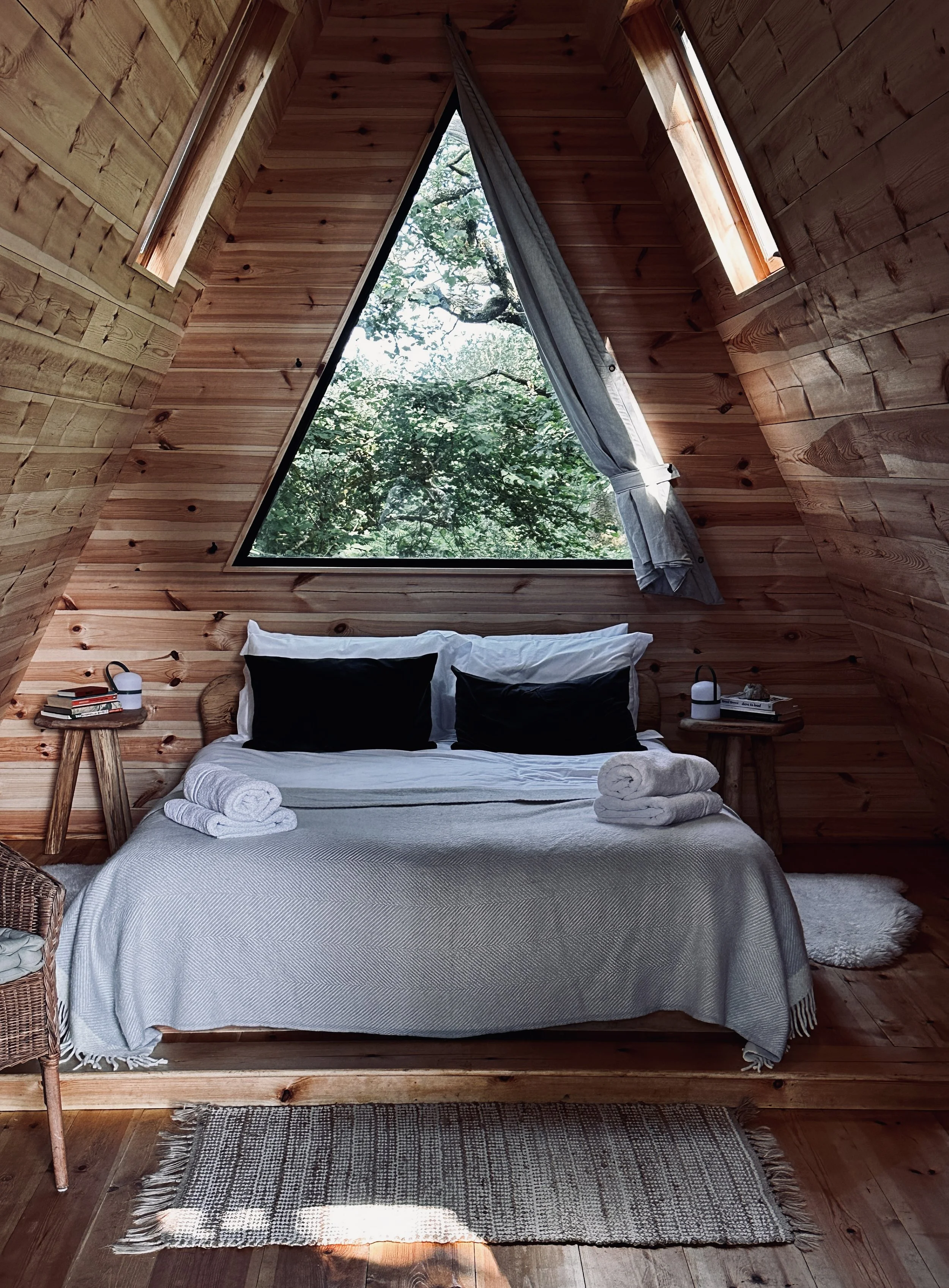 Cozy bedroom with wooden walls and ceiling, featuring a large triangular window showing green tree leaves. The bed has white sheets, black pillows, and neatly folded white towels on top. Two small side tables hold lamps and books, and a wicker chair 
