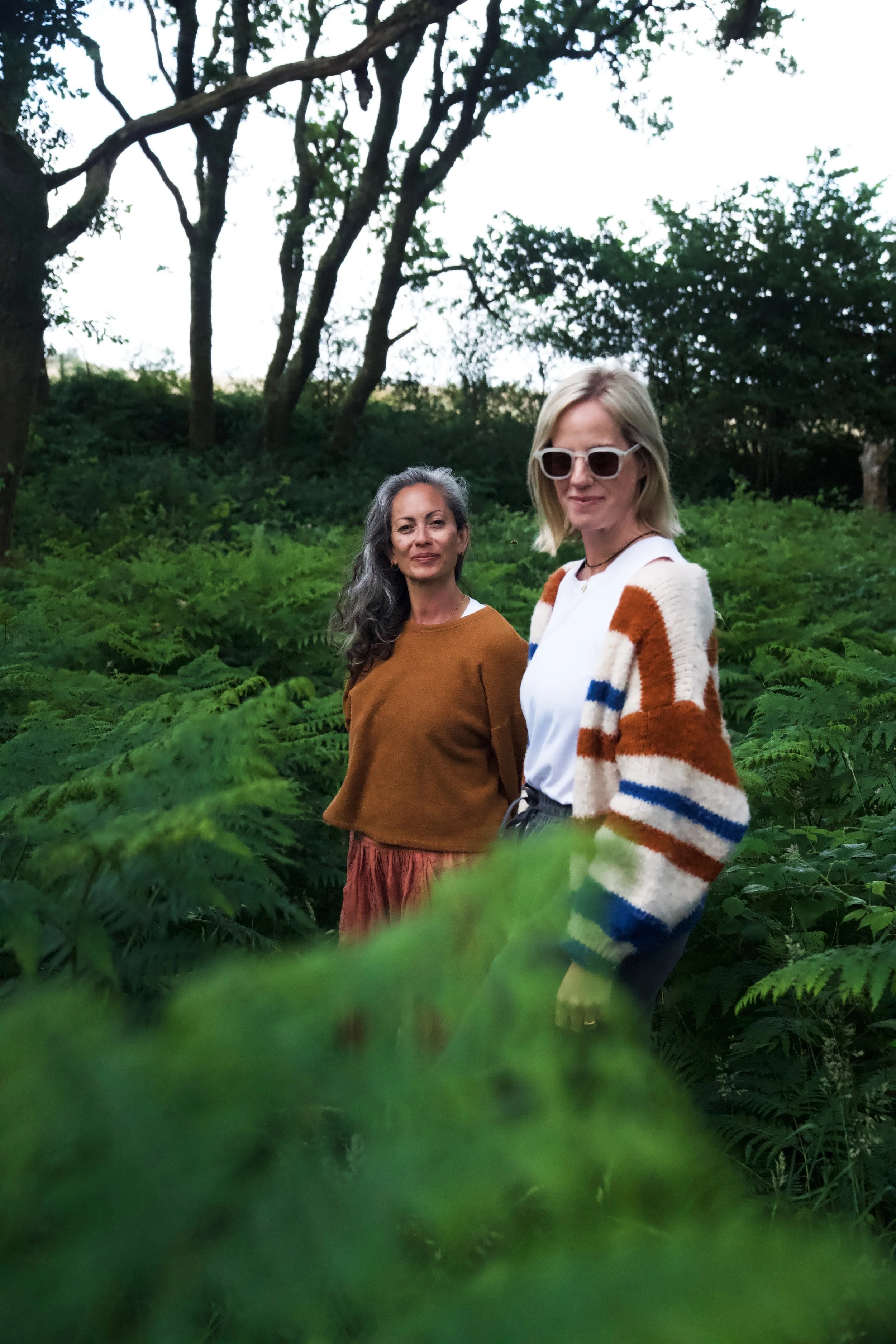 Two women standing amid lush green ferns and trees in a forested area during daylight.