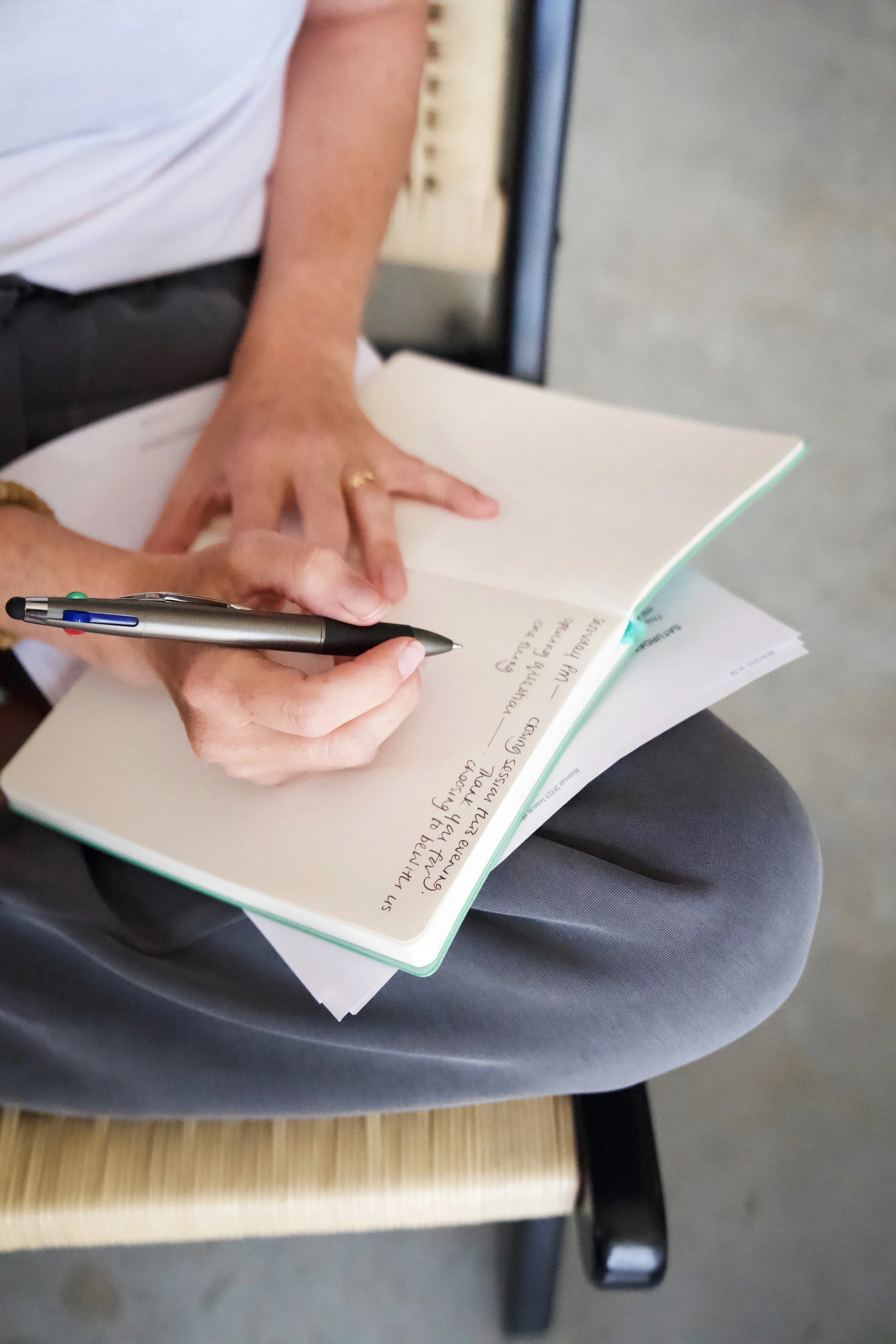Person seated, writing notes in a notebook with a black pen, other papers beneath the notebook.