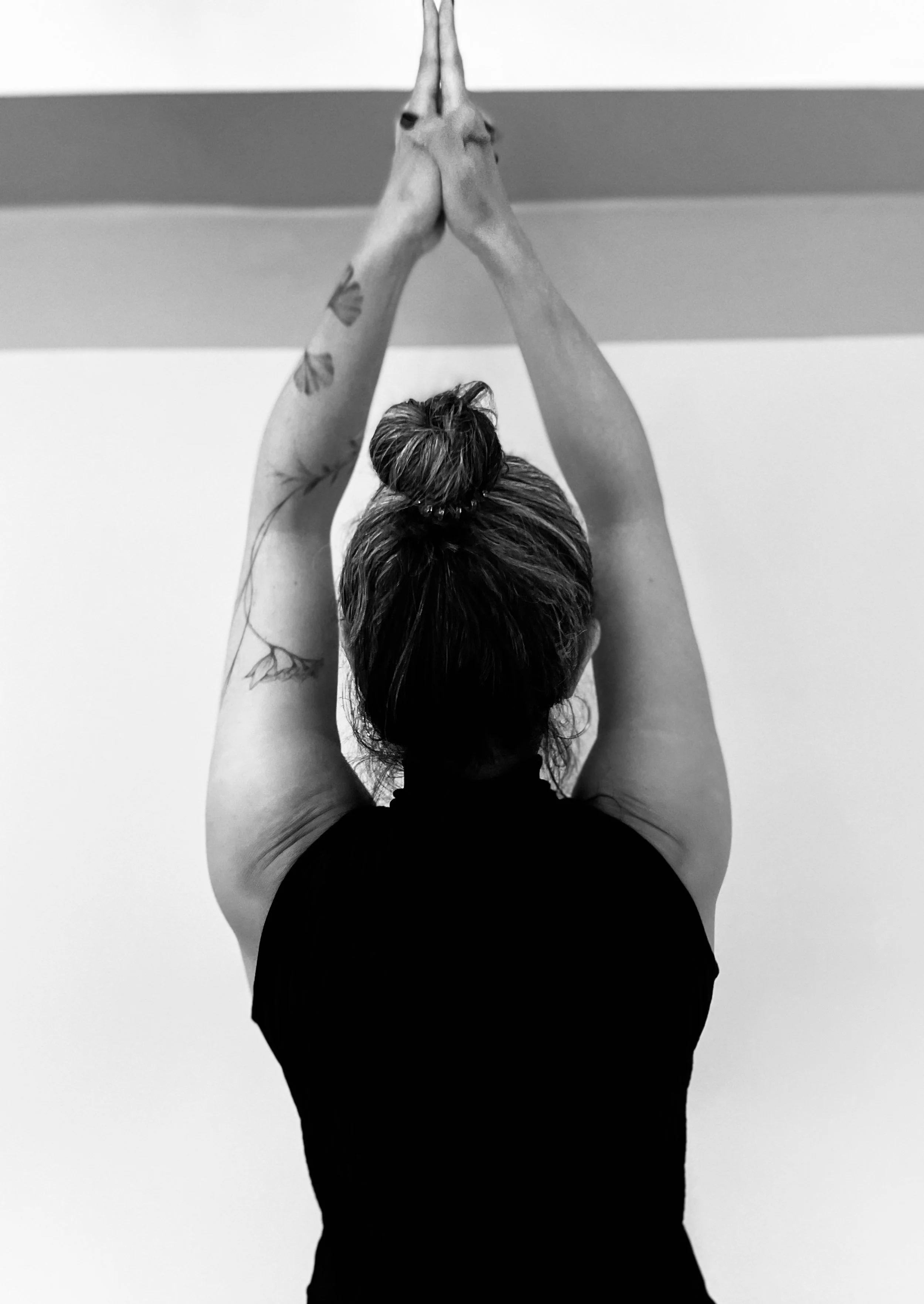A person with their back to the camera, arms raised above their head with palms pressed together in a yoga pose, wearing a black top and having a top bun hairstyle with tattoos on their arms.
