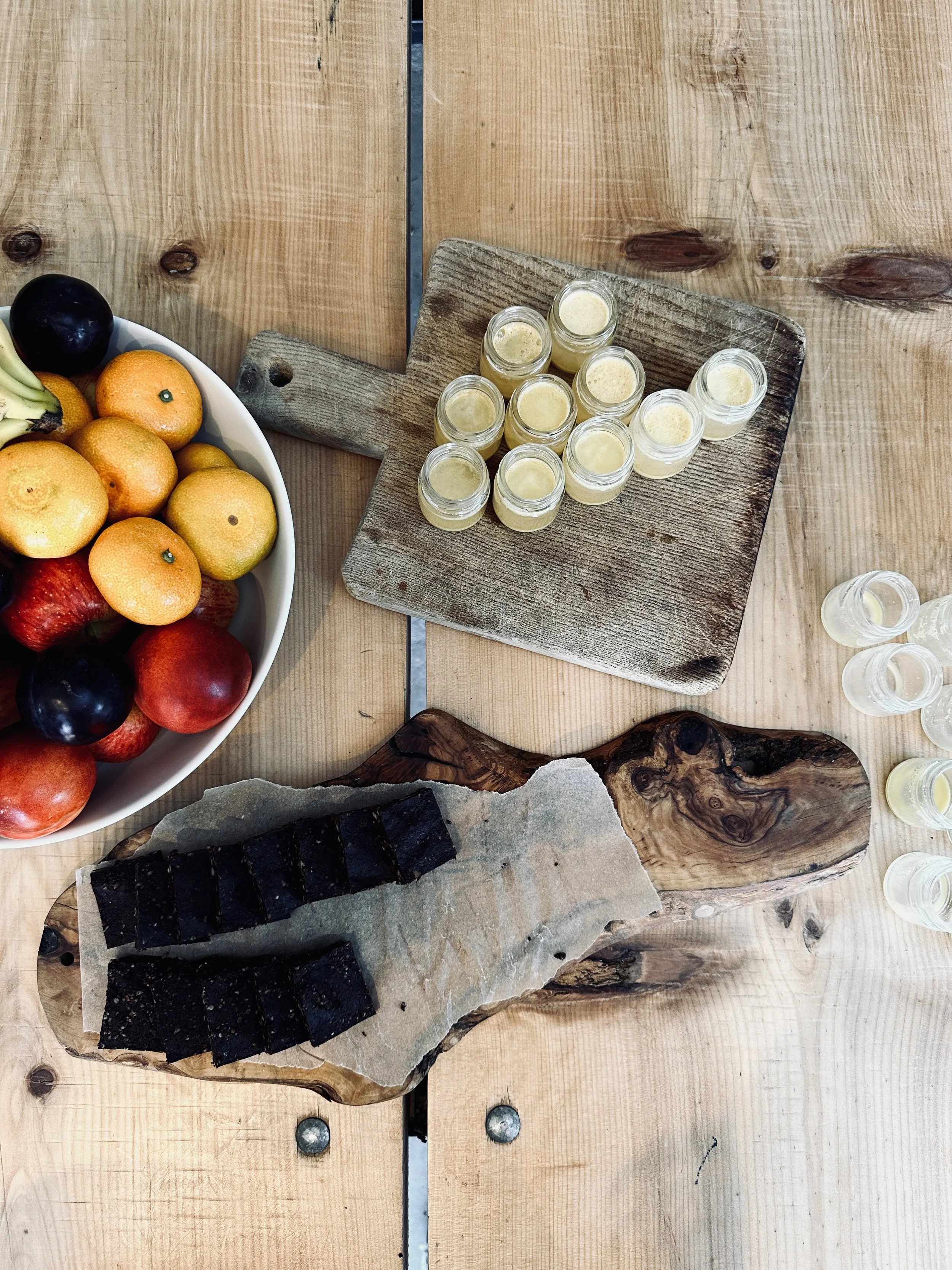 A wooden table with a bowl of mixed fruits, a cutting board with small jars of a yellow beverage, and a wooden platter with sliced dark-colored food.