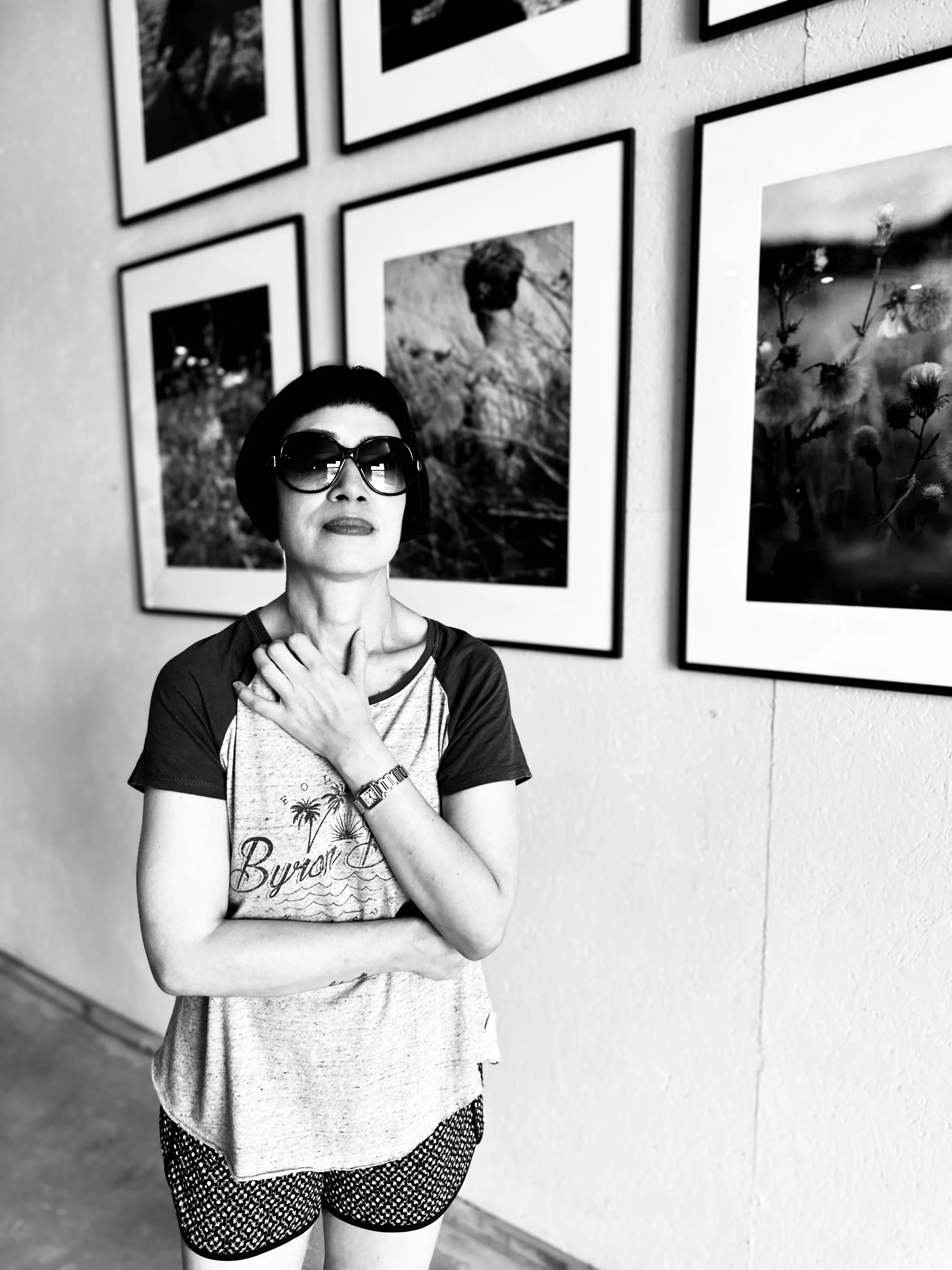 A woman wearing large sunglasses, a short-sleeved t-shirt with a palm tree and sunset print, and patterned shorts, standing in front of framed pictures on a wall.