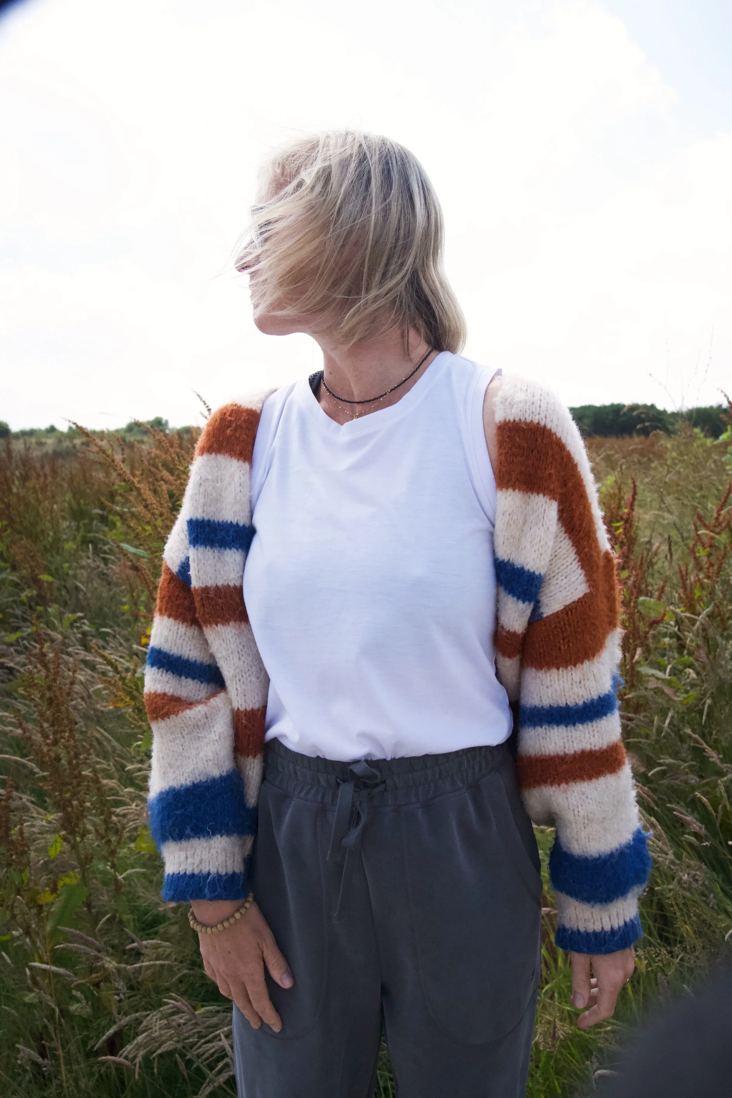 A woman with shoulder-length blonde hair, wearing a white tank top and a colorful striped cardigan in orange, white, blue, and beige, standing in a field of tall grass with an overcast sky.