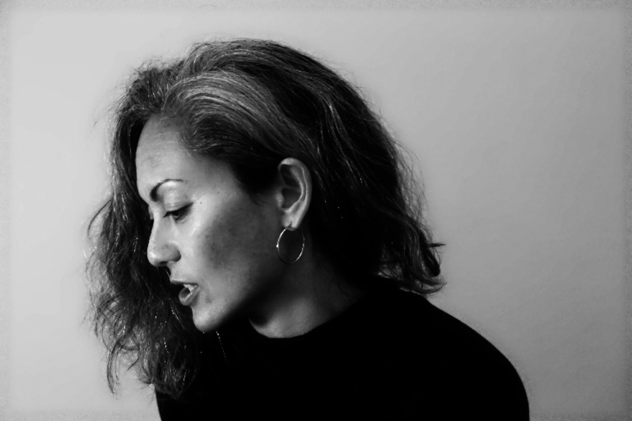 A black and white close-up portrait of a woman with shoulder-length wavy hair, wearing a hoop earring, with her eyes closed and head slightly tilted downward.