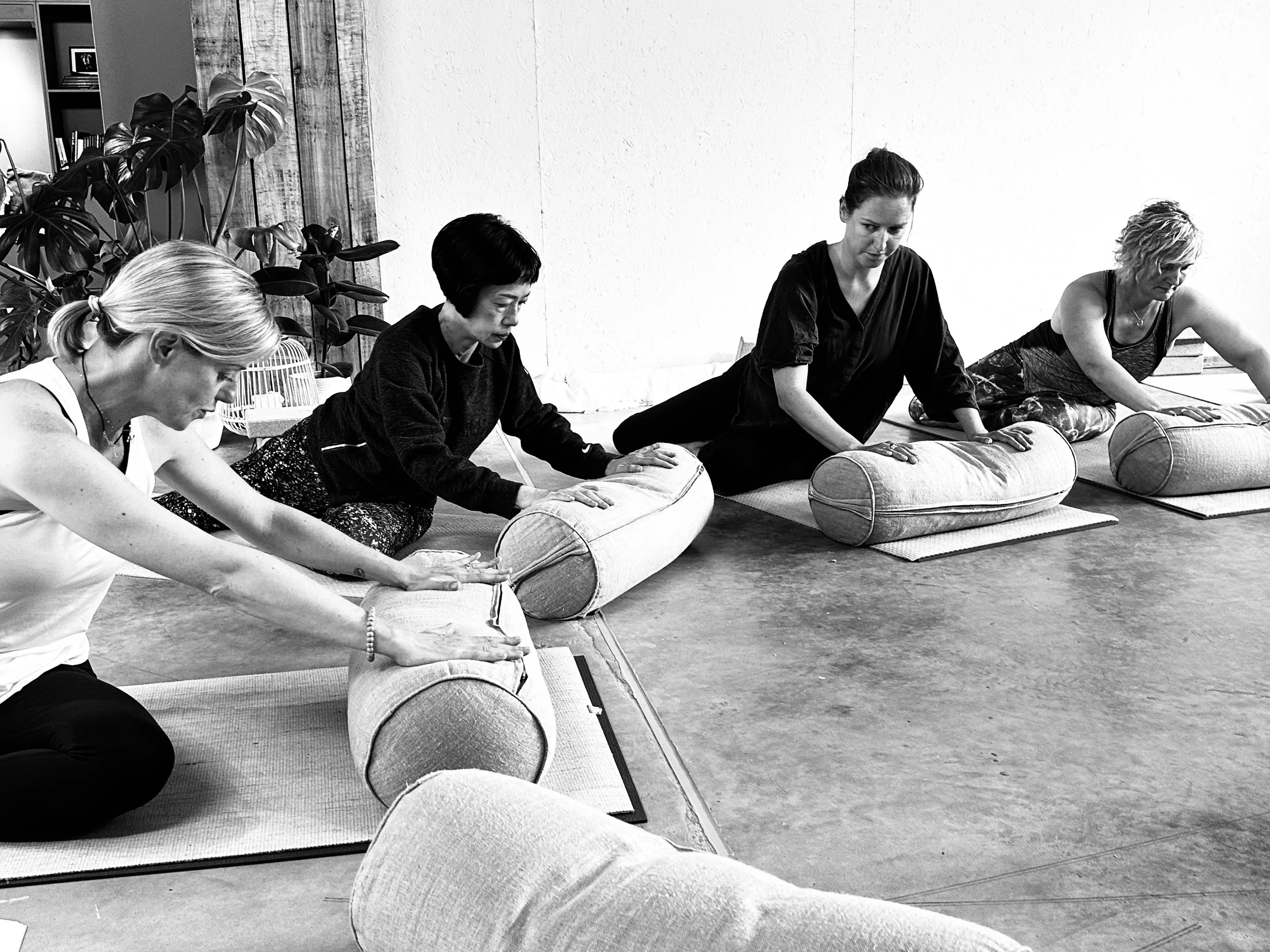 Four women participating in a yoga class, sitting on mats and using cylindrical bolsters for support during a stretching pose.