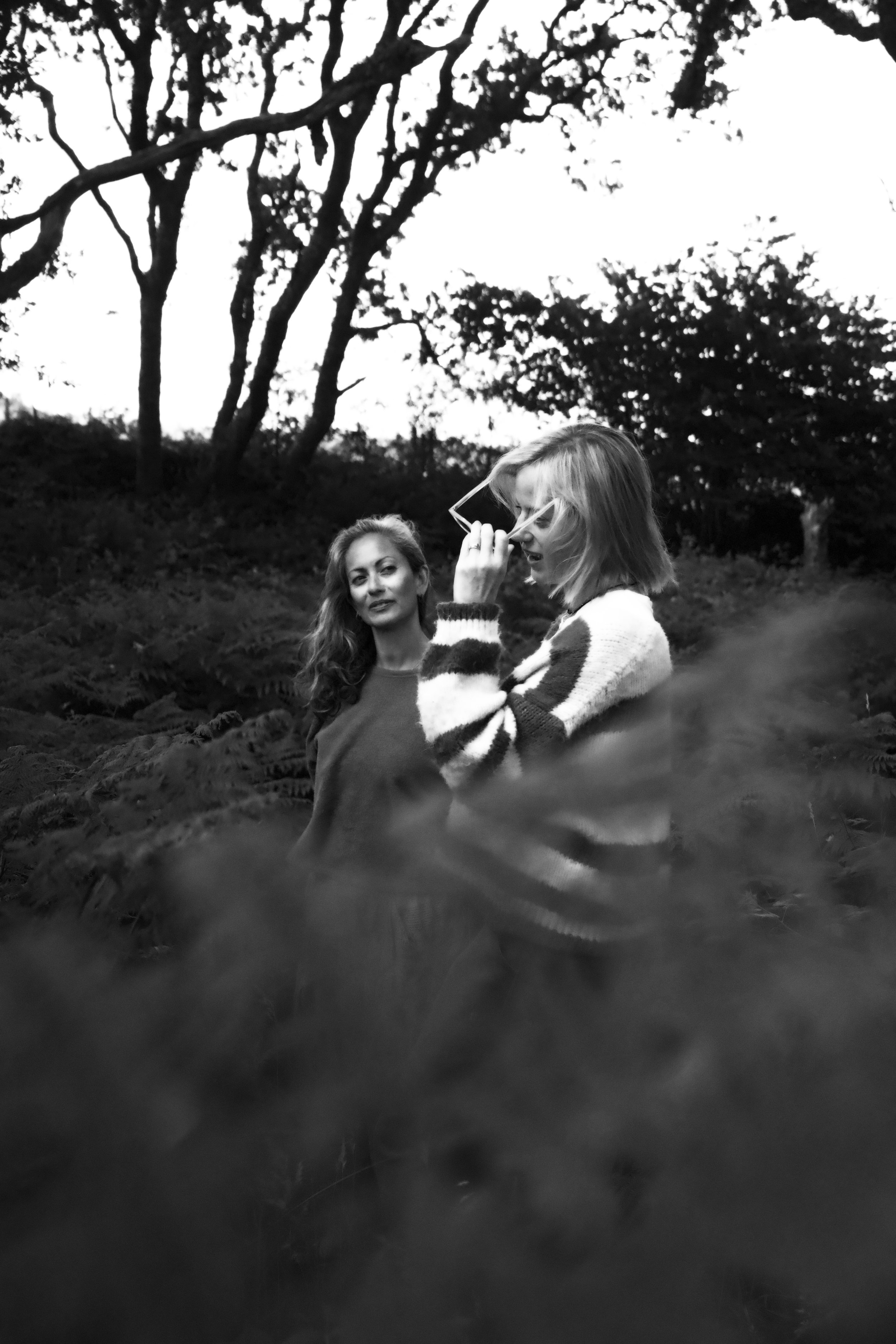 Black and white photo of two women outdoors, one with curly hair looking at the camera, the other with straight hair holding glasses near her face, surrounded by trees and foliage.