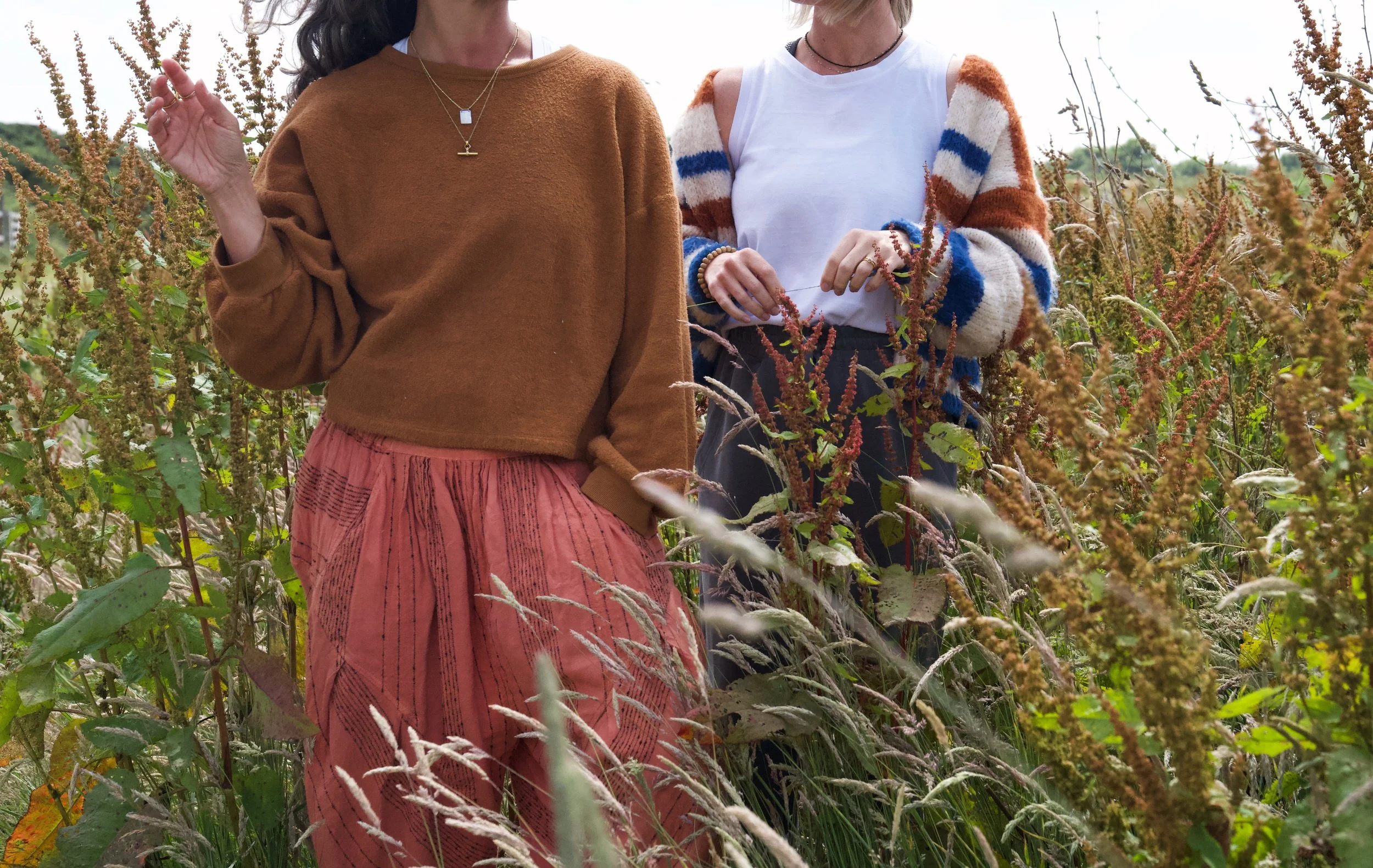 Two women standing in a field of tall grass and plants during daytime, dressed in casual clothing with necklaces, side by side with one woman raising a hand and the other holding a plant.