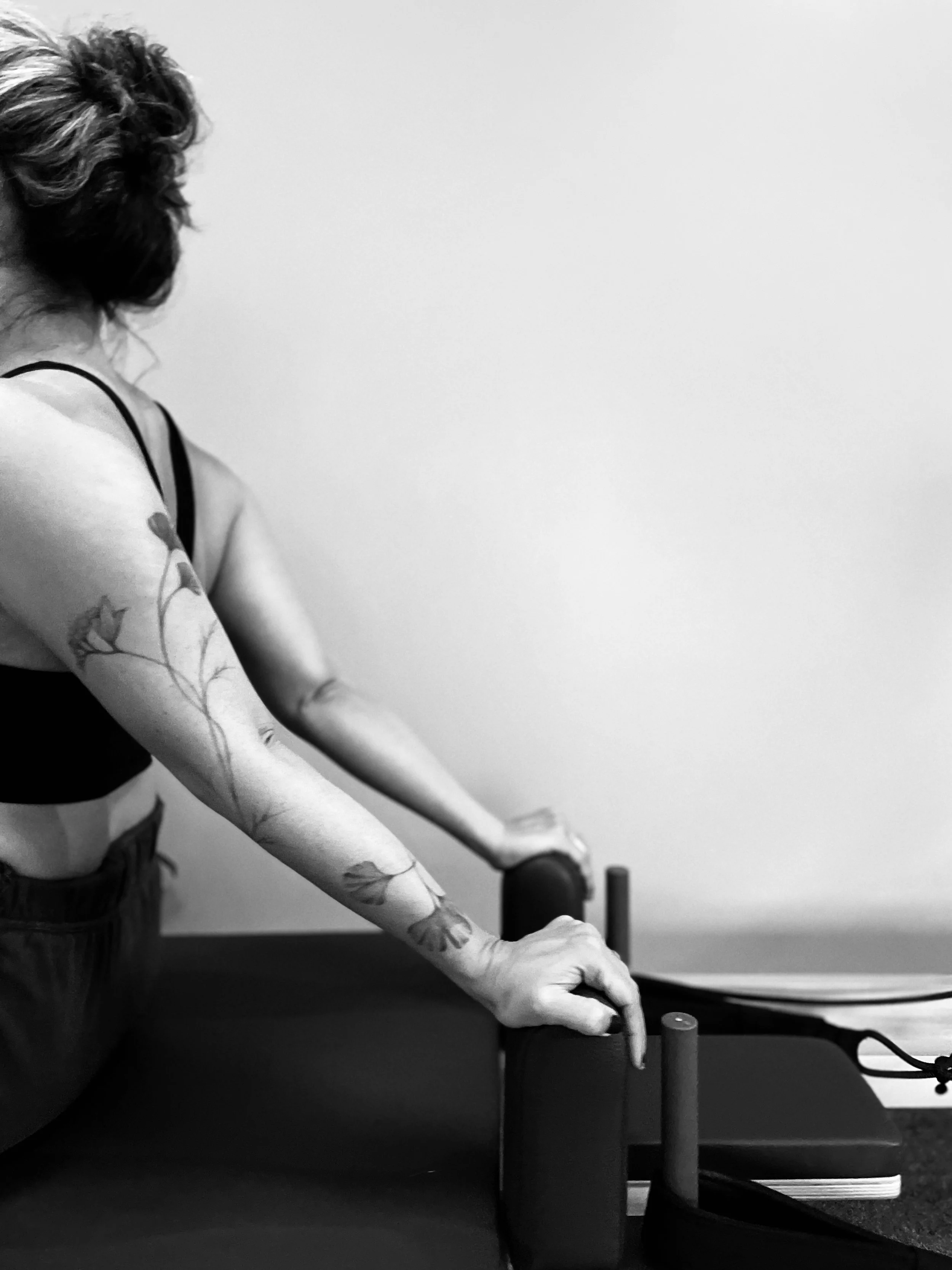 A woman with tattoos on her arm, wearing a black sports bra, is doing a push-up in a room with a plain wall. There is a chair and a desk with some papers and a computer mouse on it.