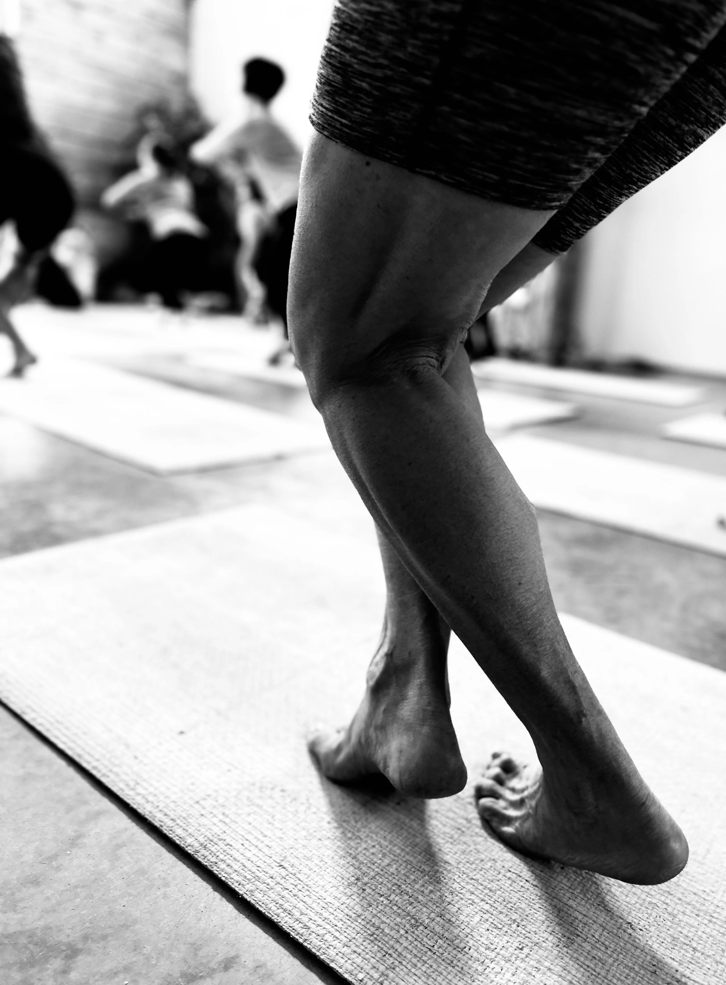 Close-up black-and-white photo of a person practicing yoga on a mat, focusing on their bent knees and feet, with other people exercising in the background.