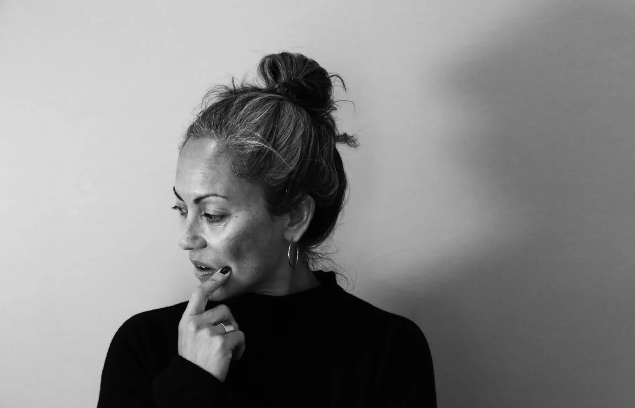 Black and white portrait of a woman with hair in a messy bun, wearing hoop earrings, with her hand on her chin, looking down thoughtfully against a plain background.