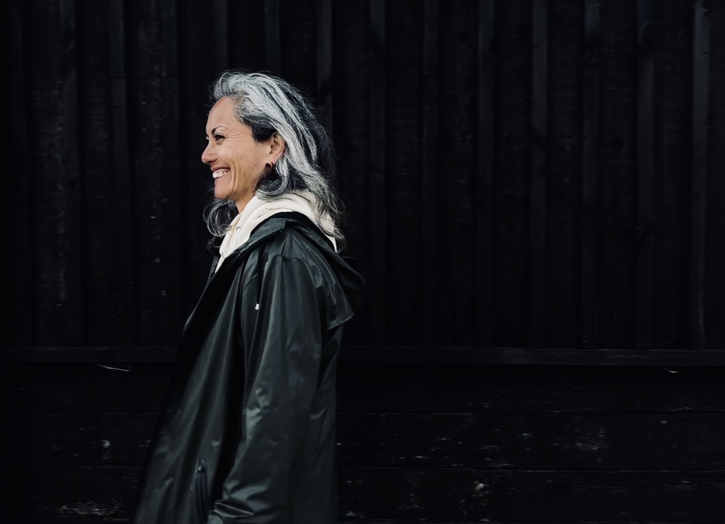 A smiling woman with gray hair, wearing a black jacket and white hoodie, walking at night against a dark wood background.