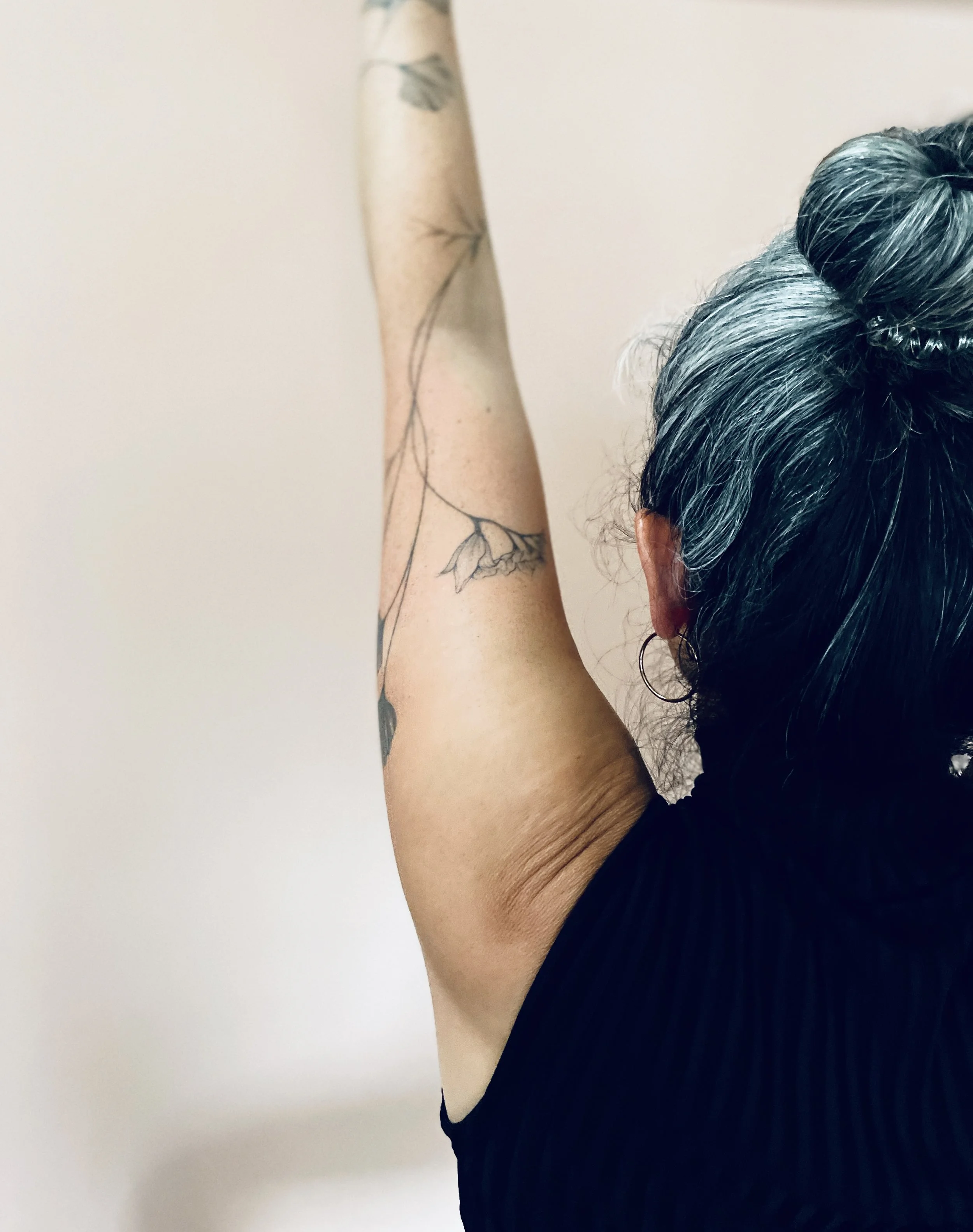 A woman with gray and black hair tied in a bun, wearing a black sleeveless shirt and a hoop earring, raising her arm to show a tattoo of a branch with leaves on her inner arm.