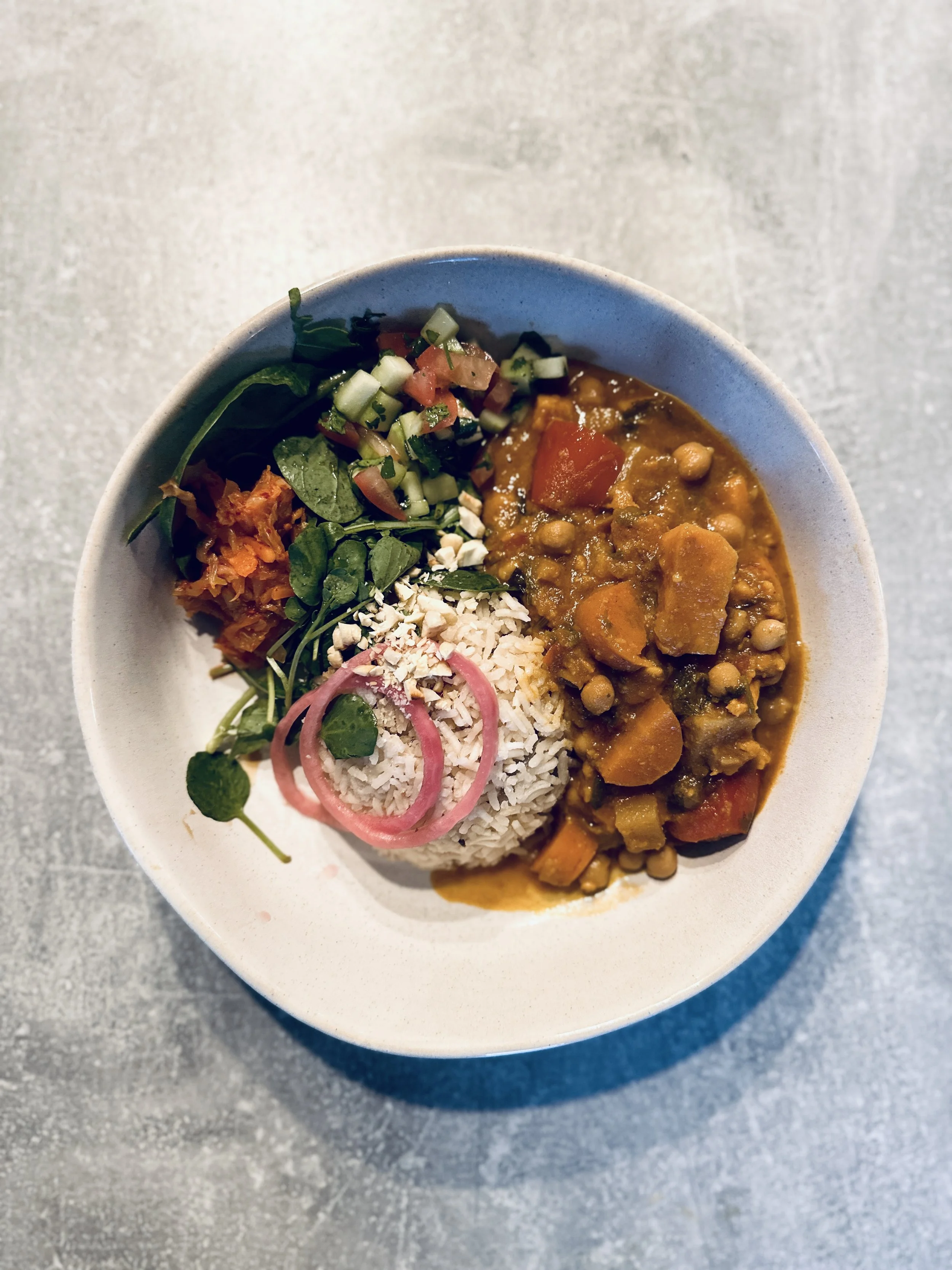 A bowl of rice and vegetable curry with side salads including chopped cucumbers, cherry tomatoes, and greens, garnished with pickled onions.