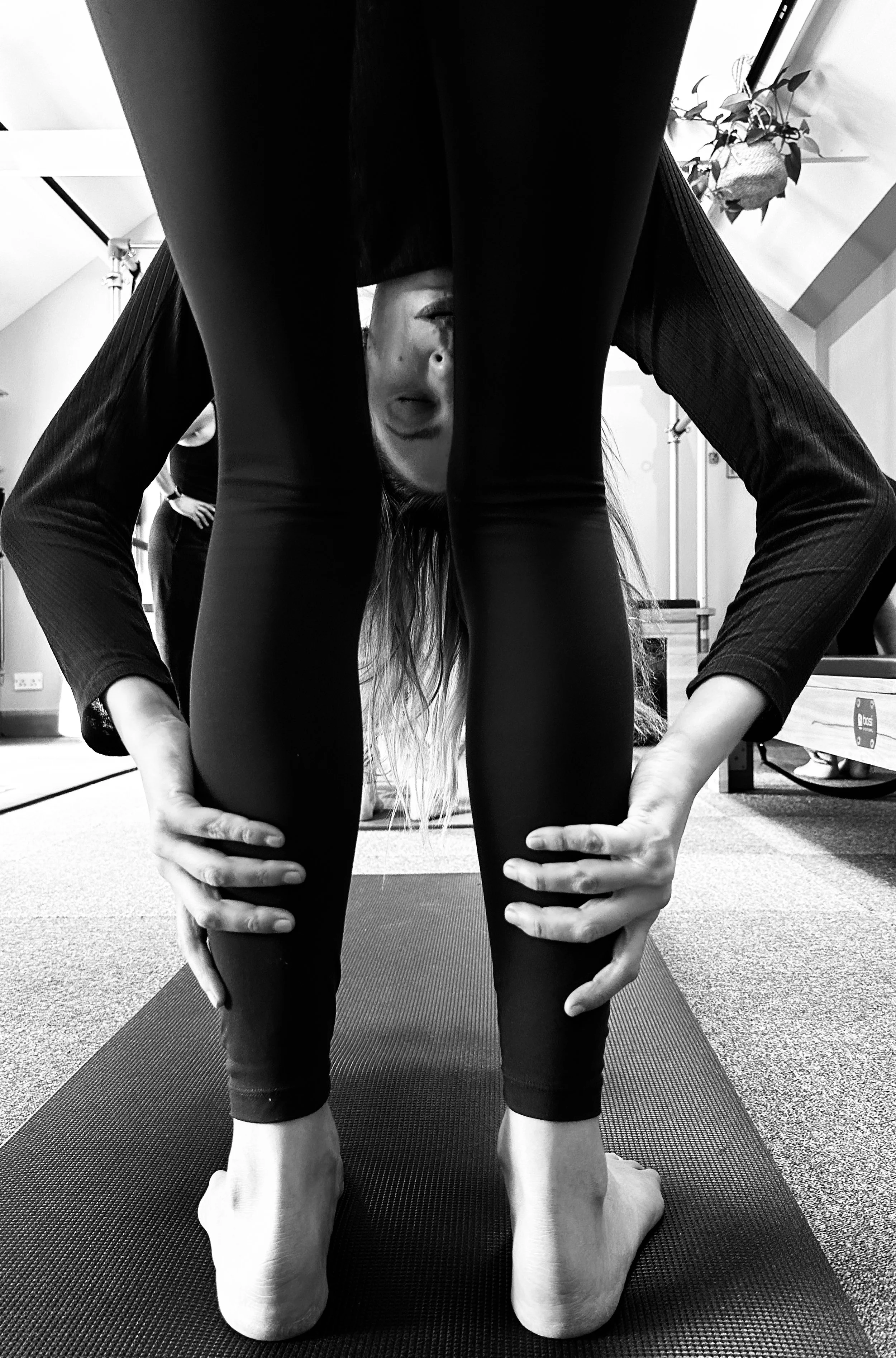 Black and white photo of a woman practicing yoga, bending forward with her head upside down between her legs, hands on her shins, on a yoga mat in a room with a plant and a lamp in the background.